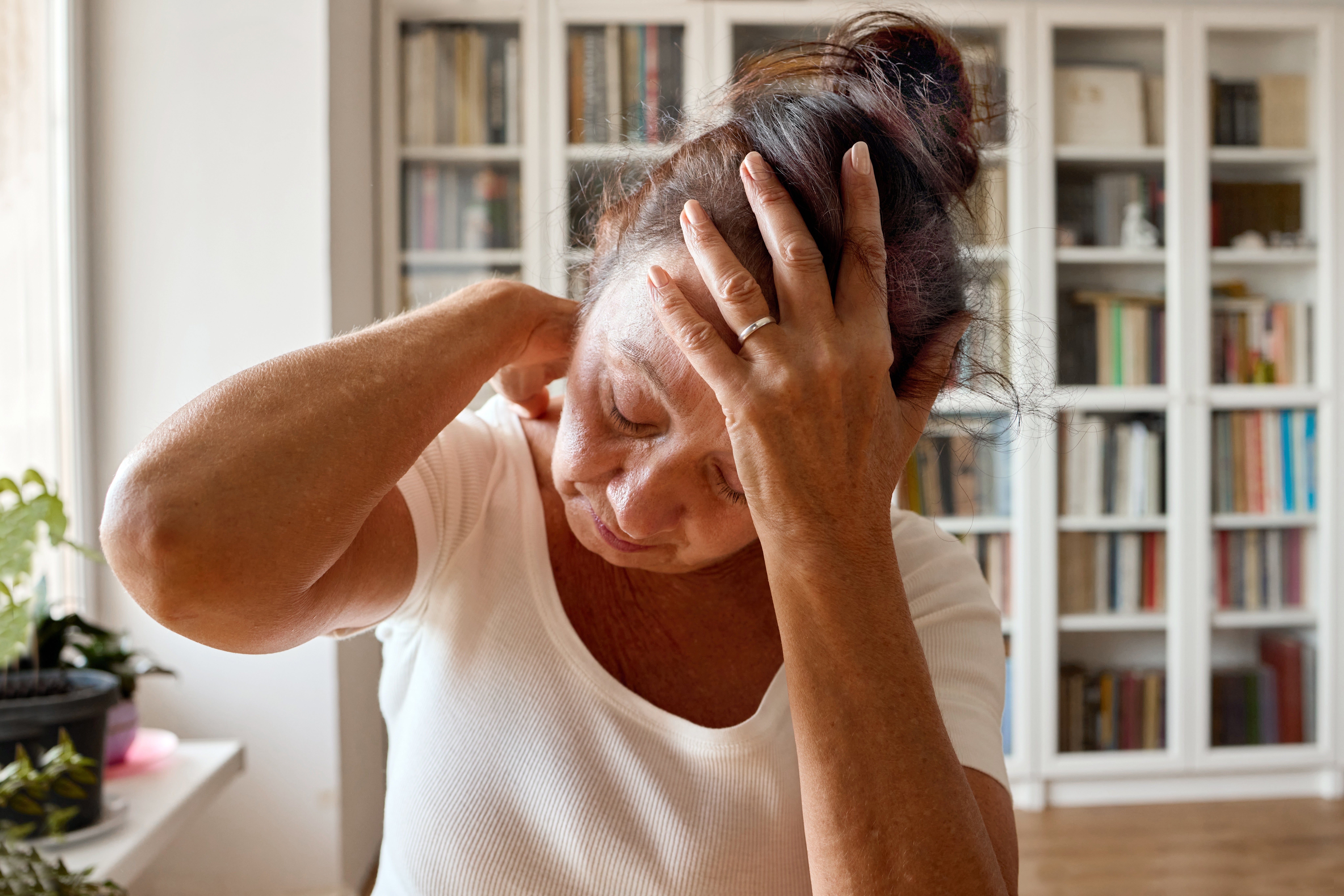 Person in a white shirt holding their neck and head, appearing stressed or in pain, in a home setting with bookshelves in the background