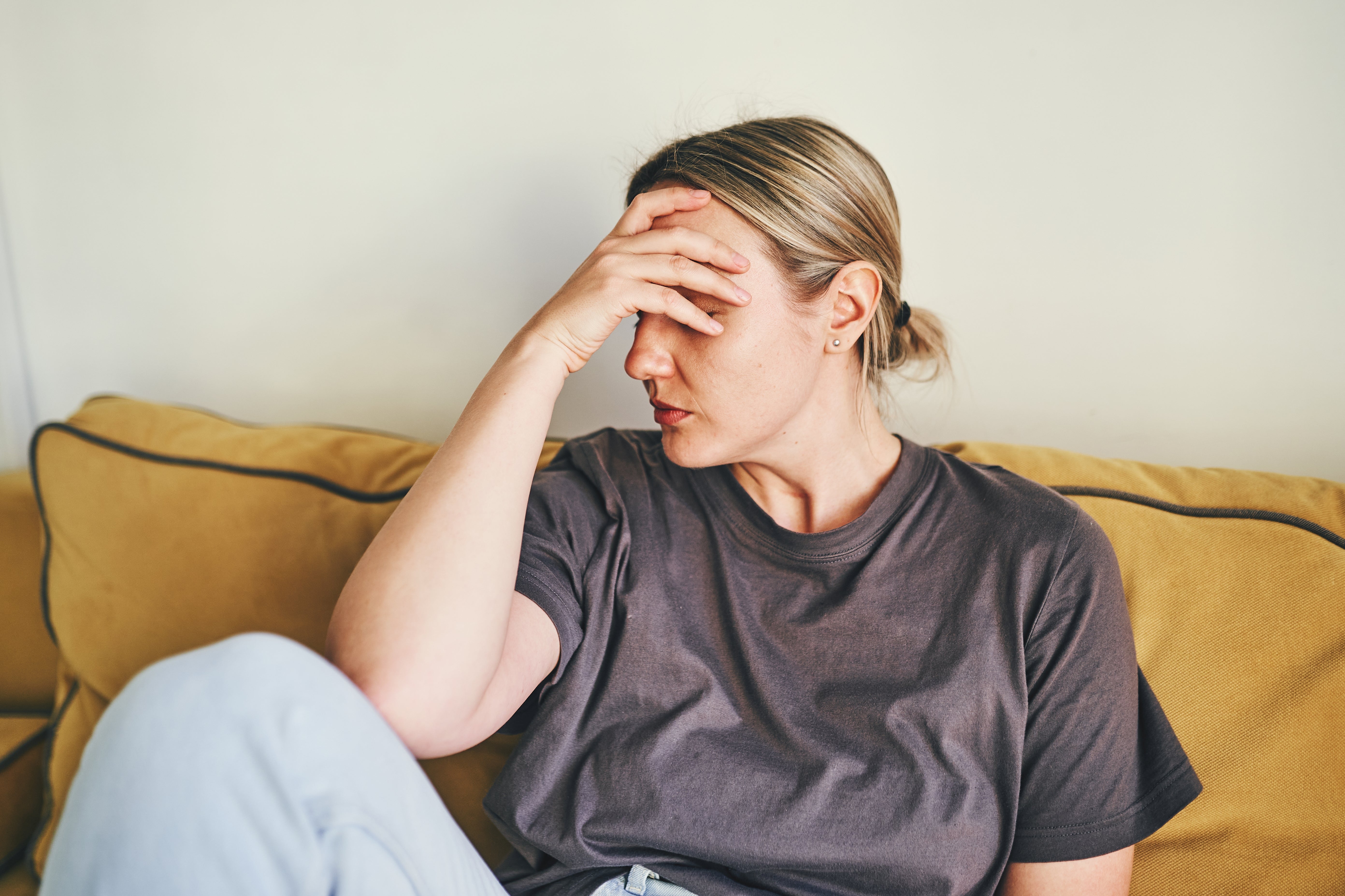 Person in casual clothing sitting on a couch, looking distressed, with their hand covering their face