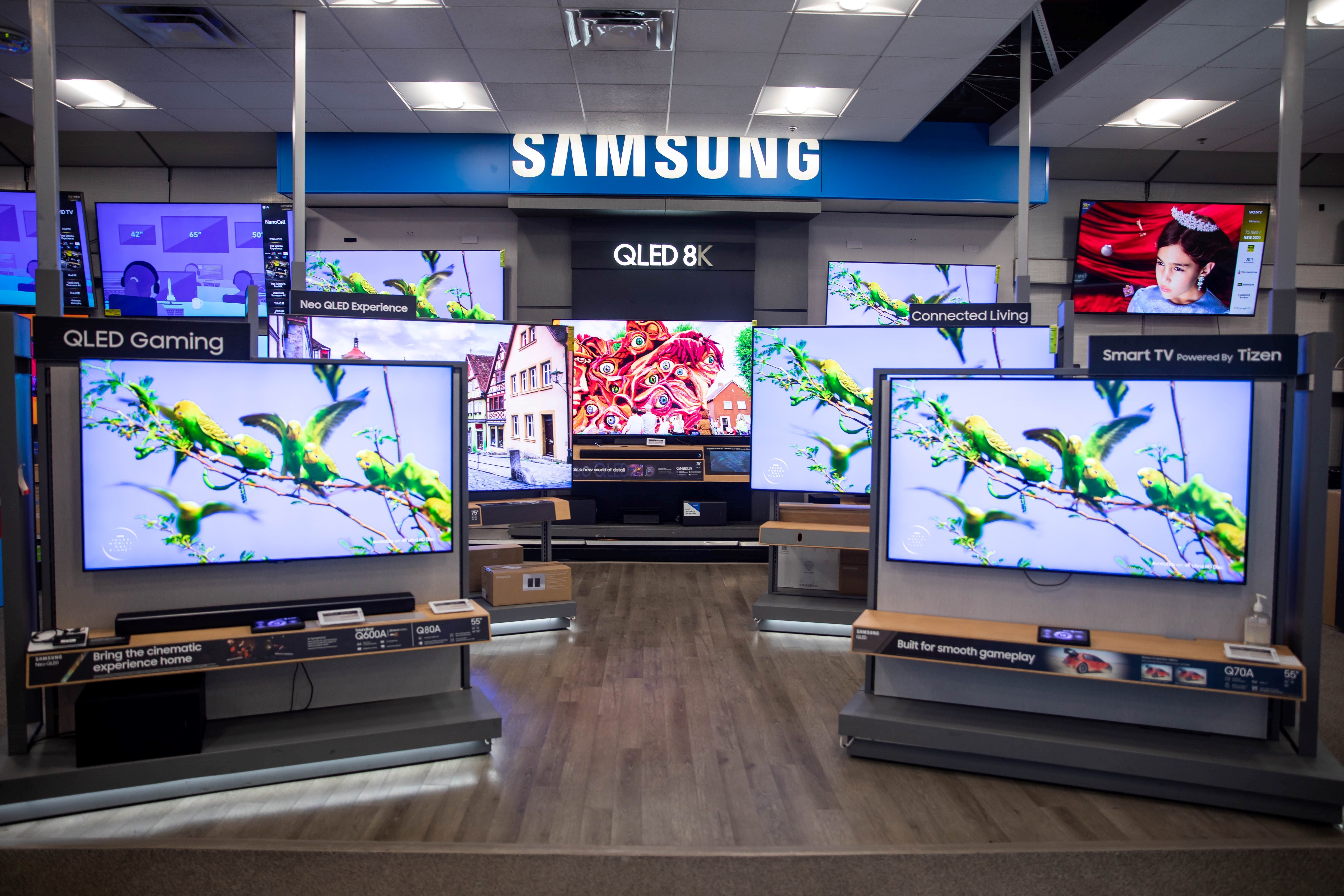 Samsung retail display featuring a range of QLED TVs with vibrant screens, showcasing models for gaming, live TV, and smart features