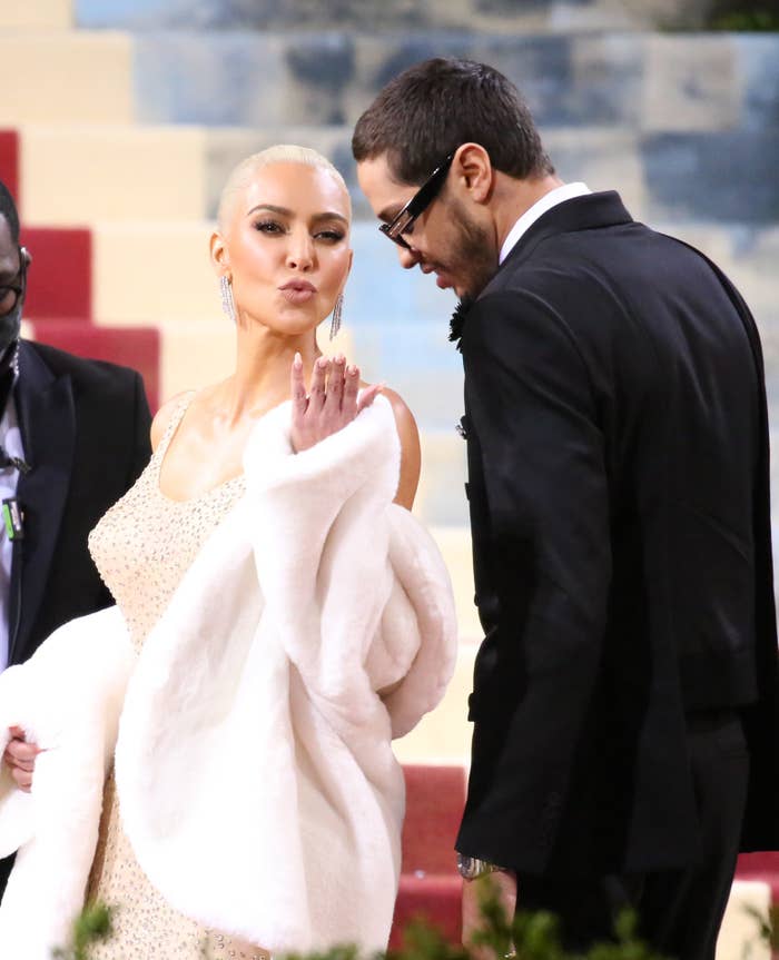 Person in elegant evening gown with a white shawl blows a kiss on a red carpet. Another person in a suit stands beside them, smiling