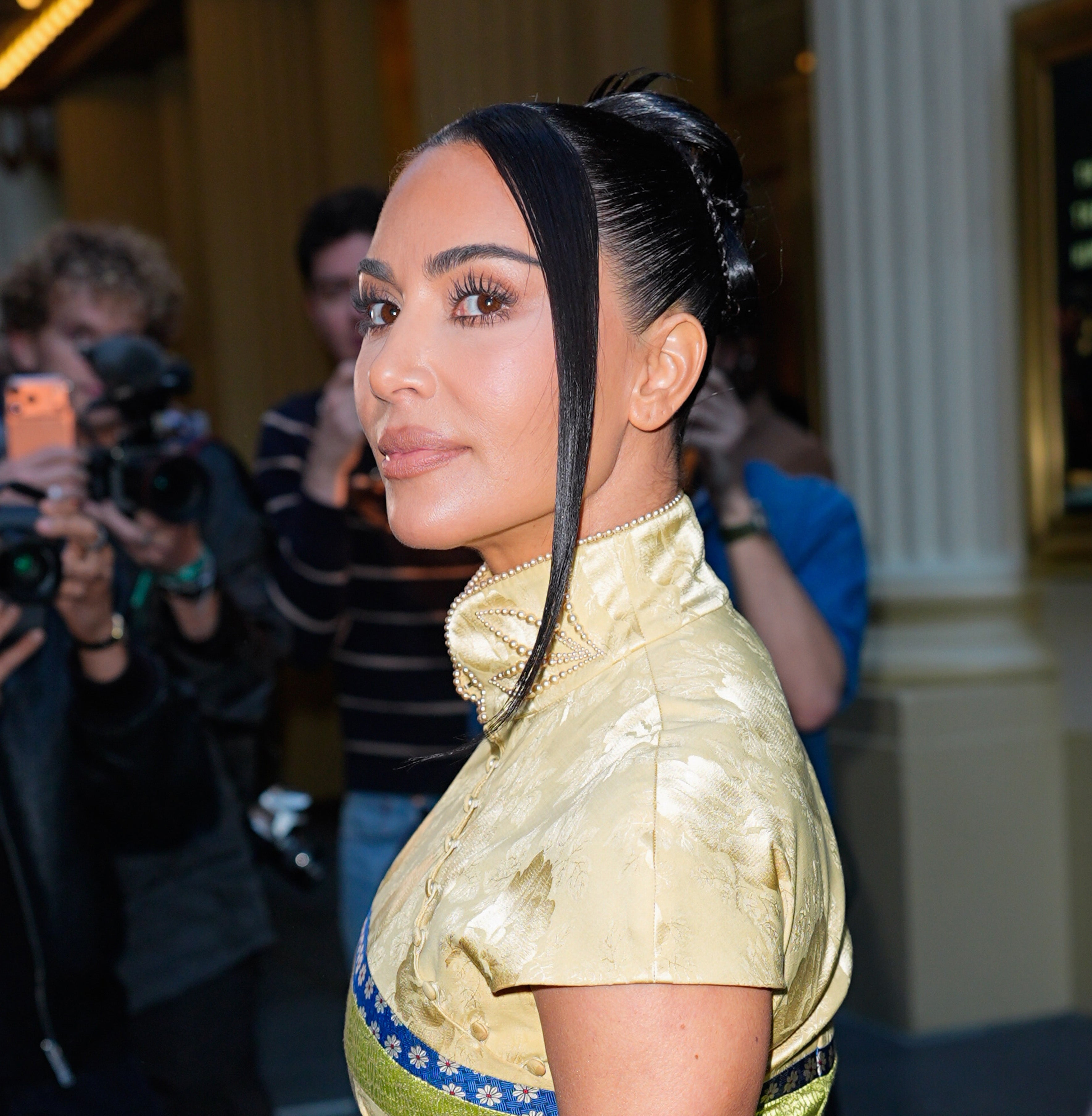 A person poses for photos wearing a stylish, high-collared dress with intricate patterns, in front of paparazzi