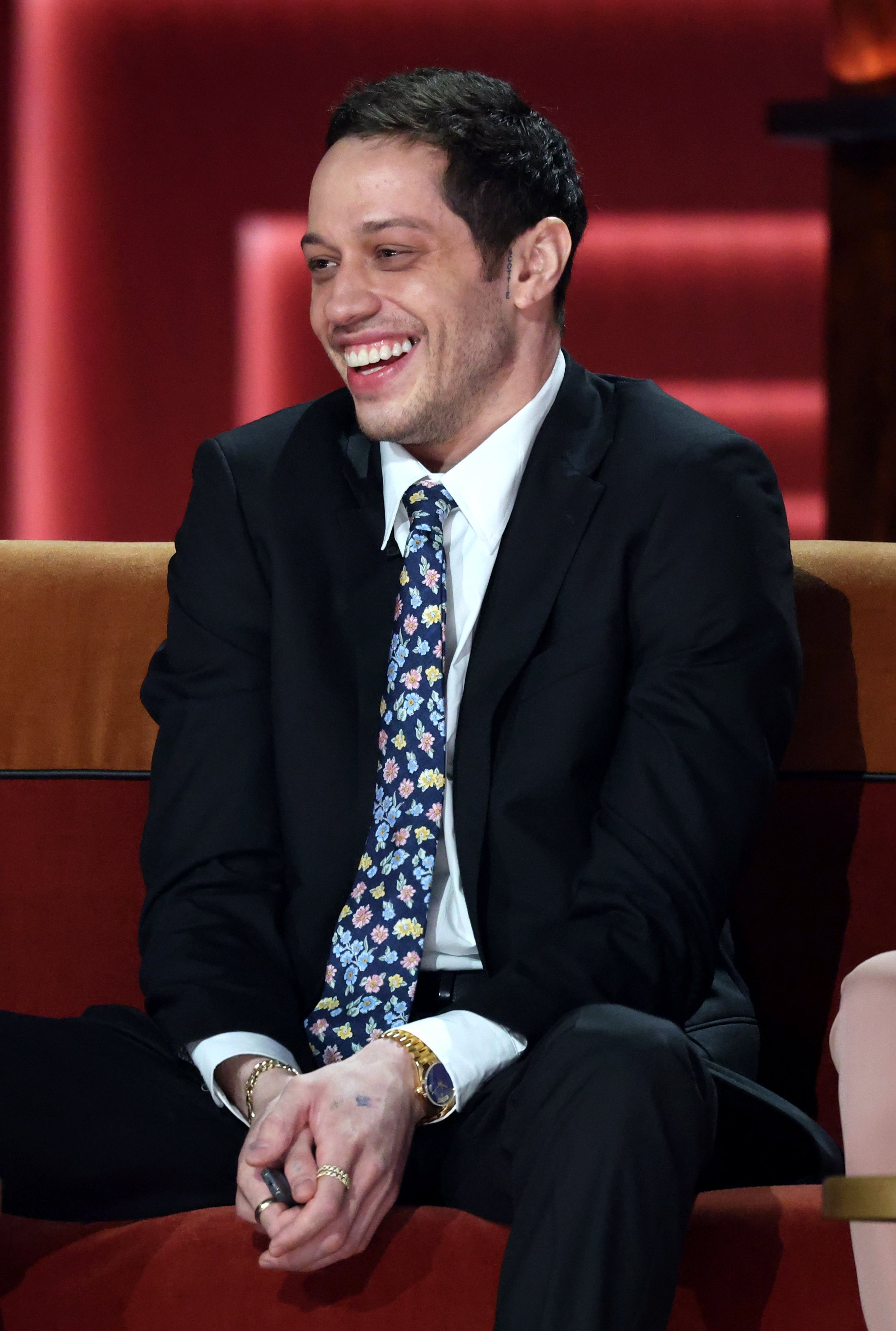 A person in a suit with a floral tie sits on a sofa, smiling, with drinks on the table in front of them