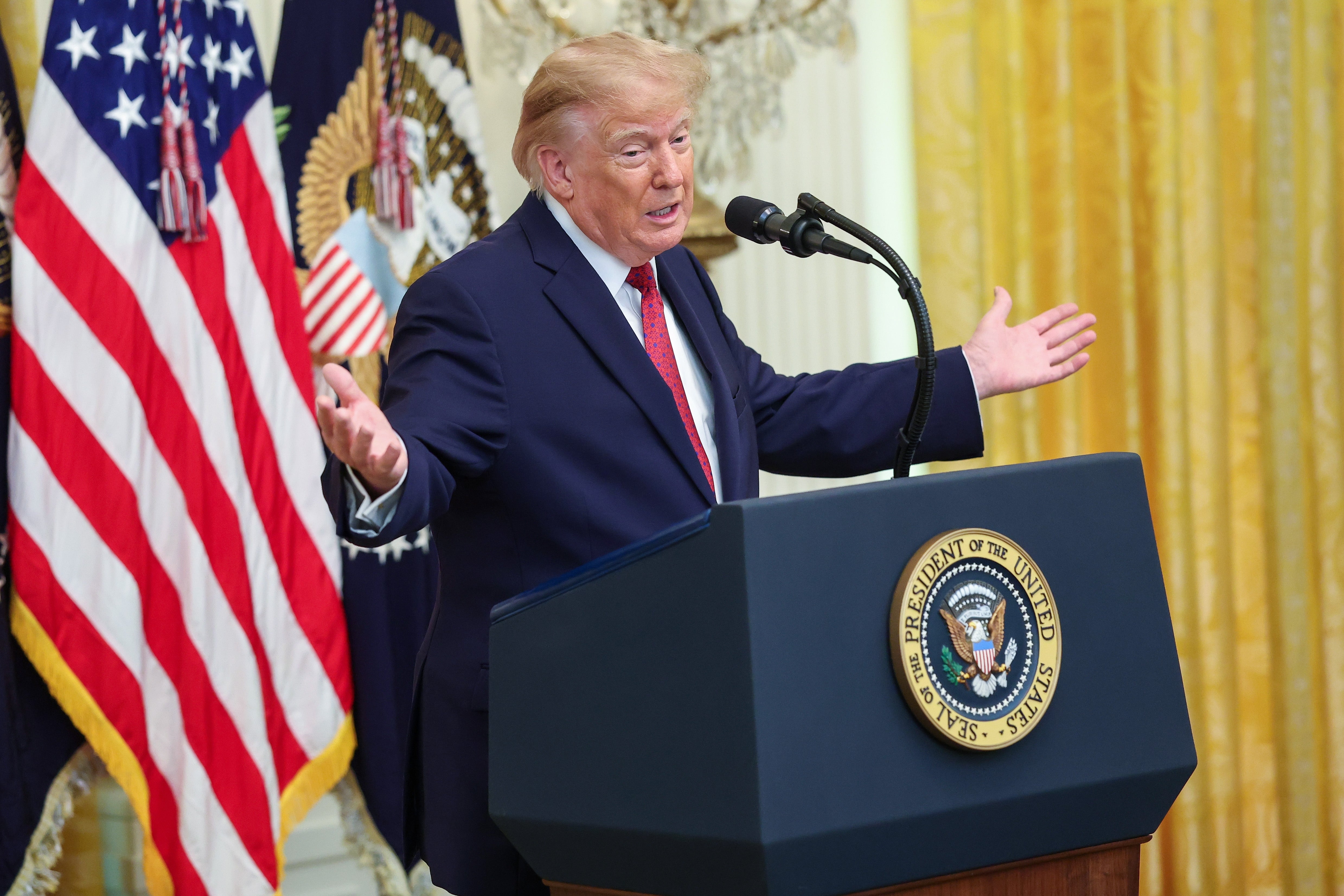 En person i dress snakker på et podium med presidentseglet, gestikulerer med åpne armer, i et utsmykket rom med flagg bak