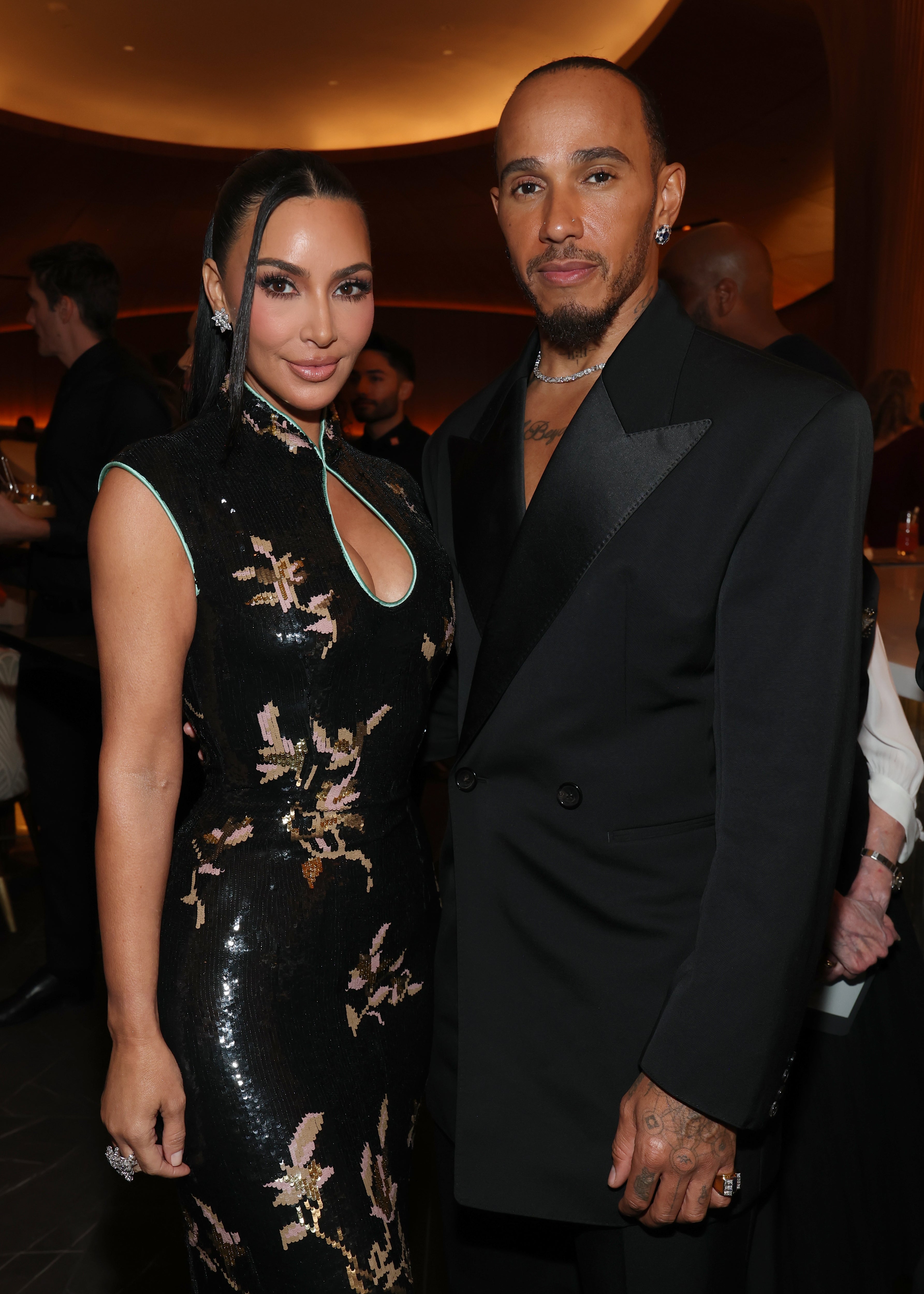 Two people pose together at an event. One is in a floral-patterned dress, and the other wears a double-breasted suit