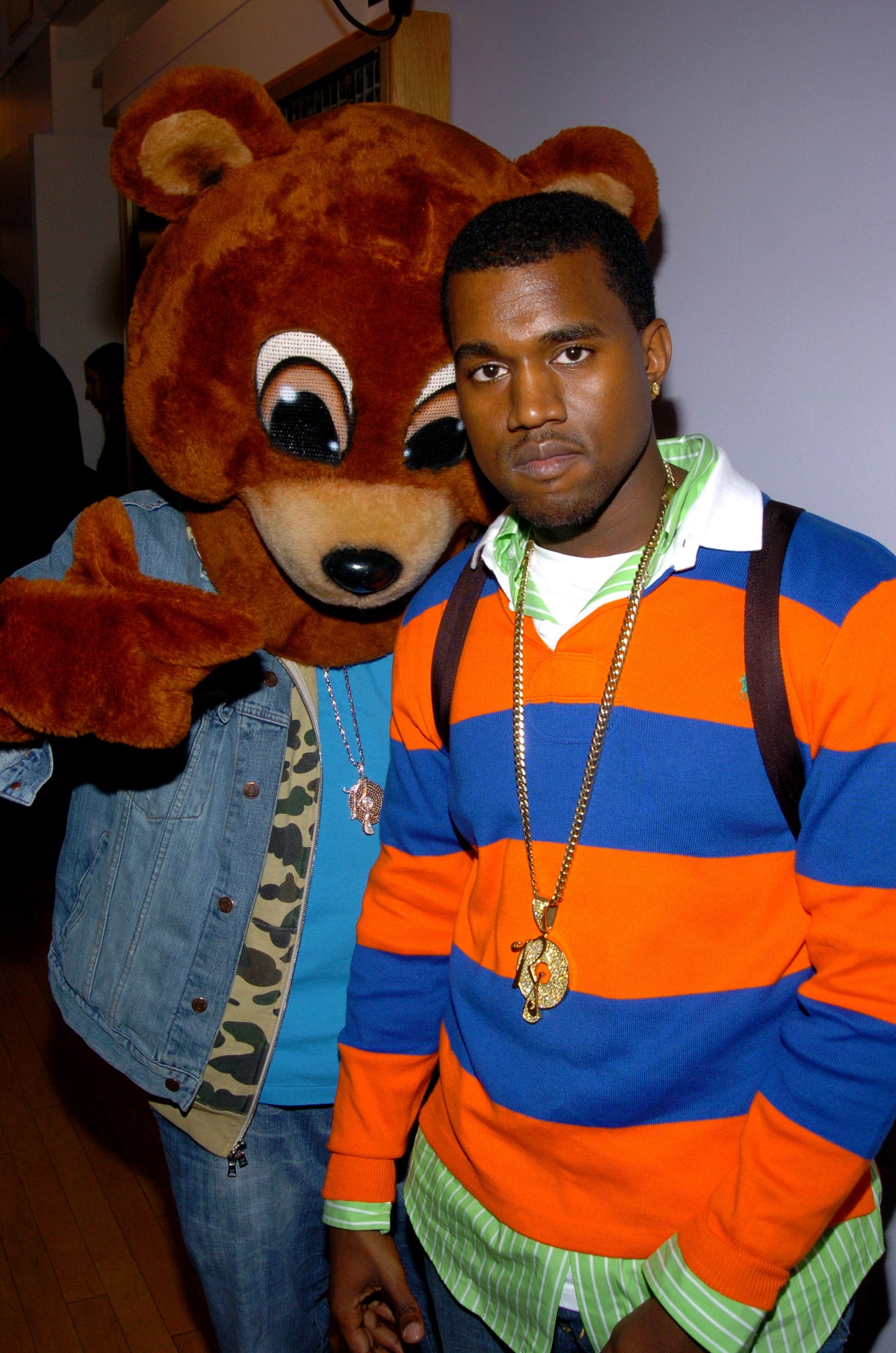 A person in a striped sweater stands next to someone in a bear mascot costume, both wearing gold necklaces