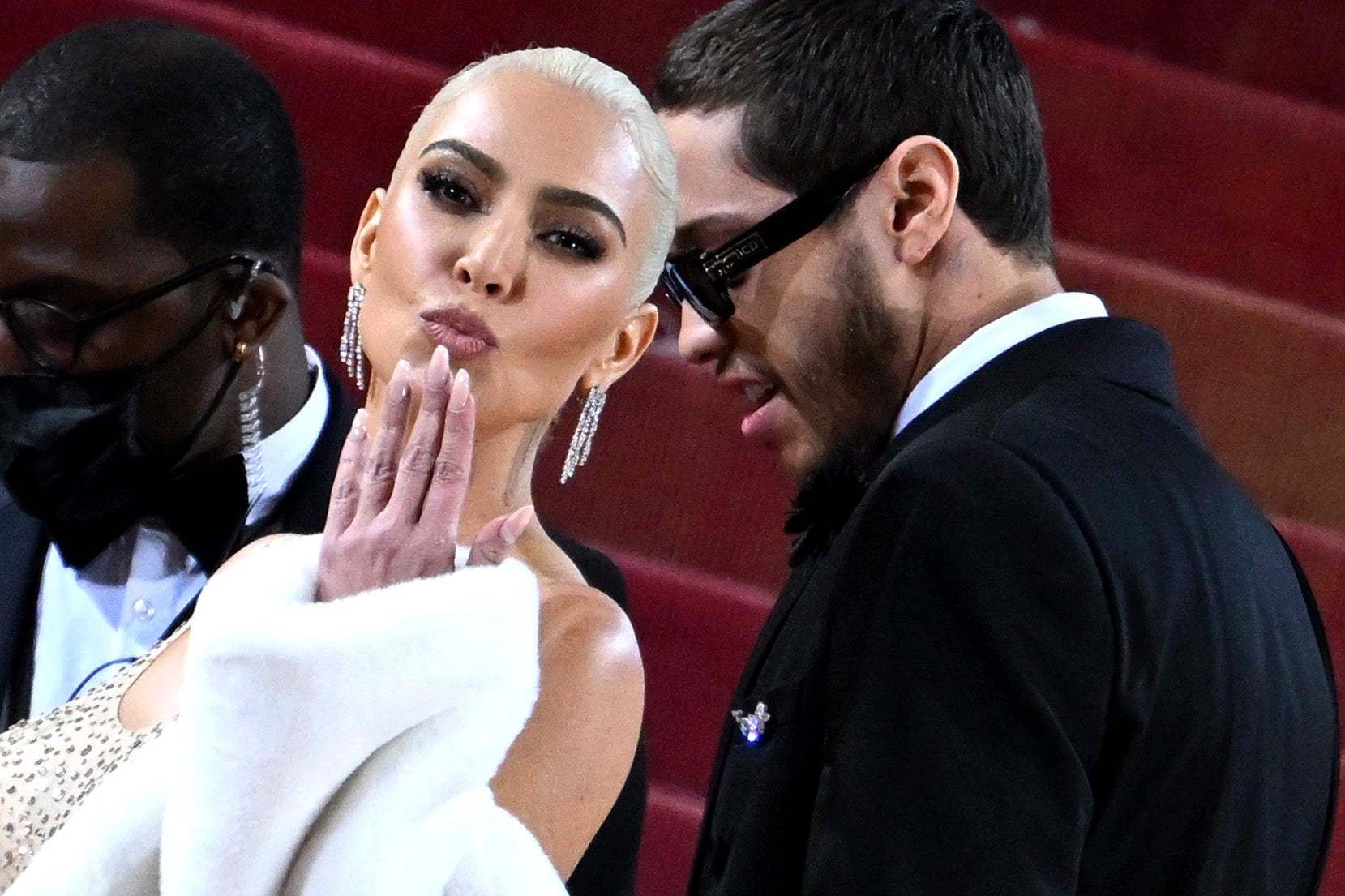 A woman in an elegant gown blows a kiss on a red carpet, accompanied by a man in a tuxedo and sunglasses