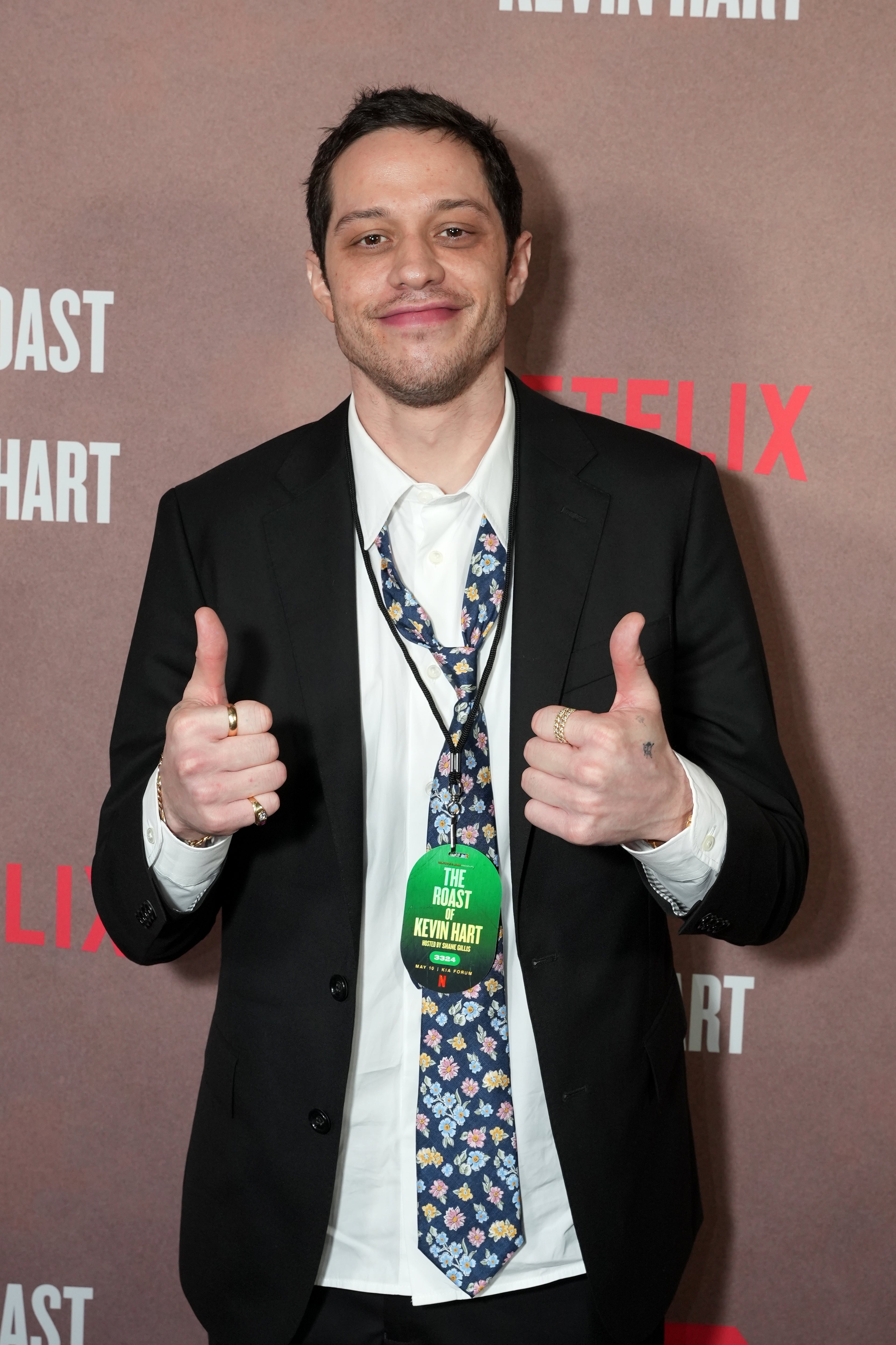 Person in a suit and floral tie gives thumbs-up on a red carpet with "Netflix" and "Roast of Kevin Hart" text in the backdrop