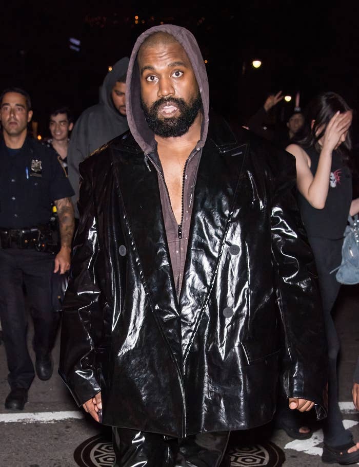 Person in a shiny oversized jacket and hoodie, walking on a street at night with people and police officers in the background
