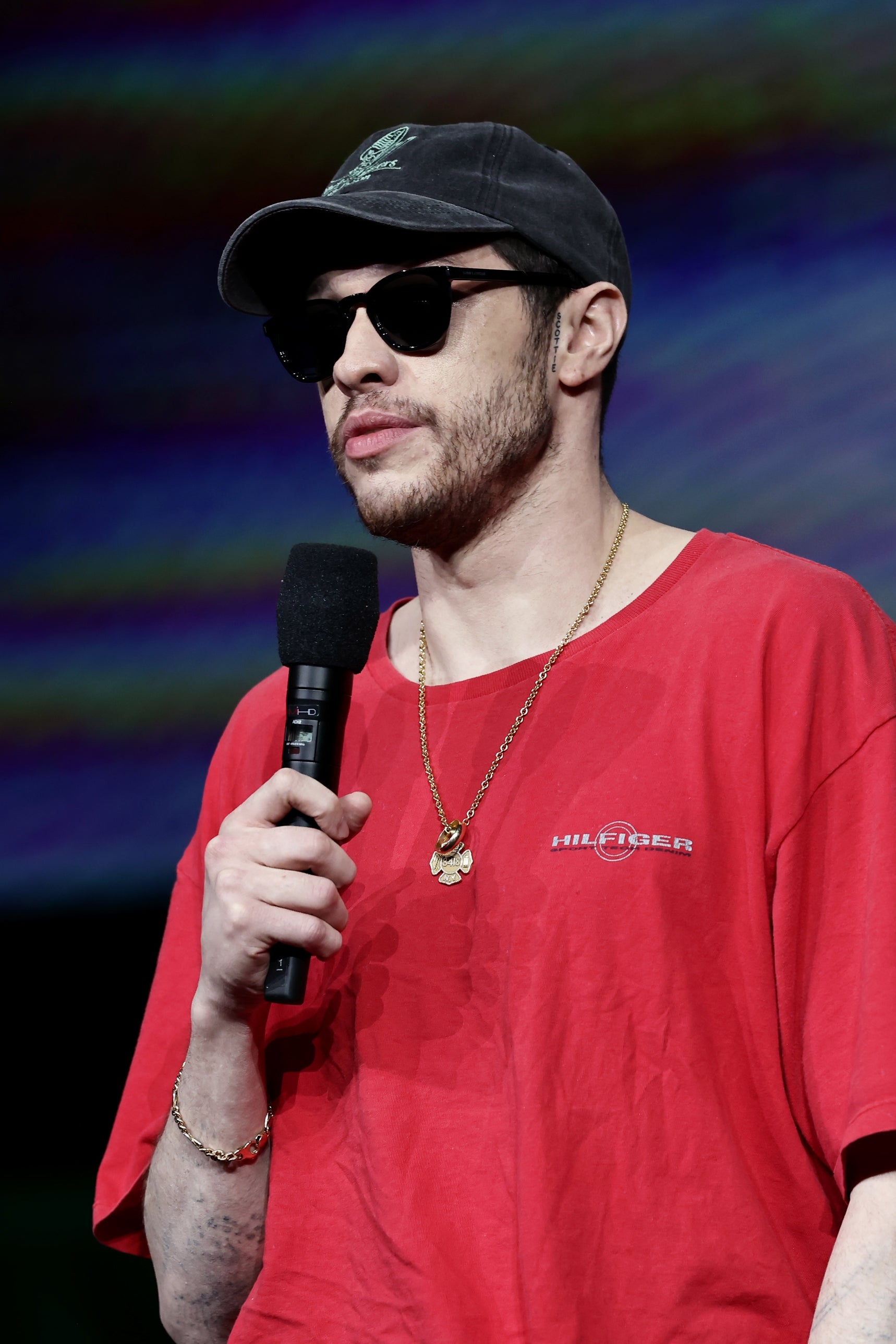 Person wearing a cap, sunglasses, and a casual red T-shirt speaks into a microphone on stage