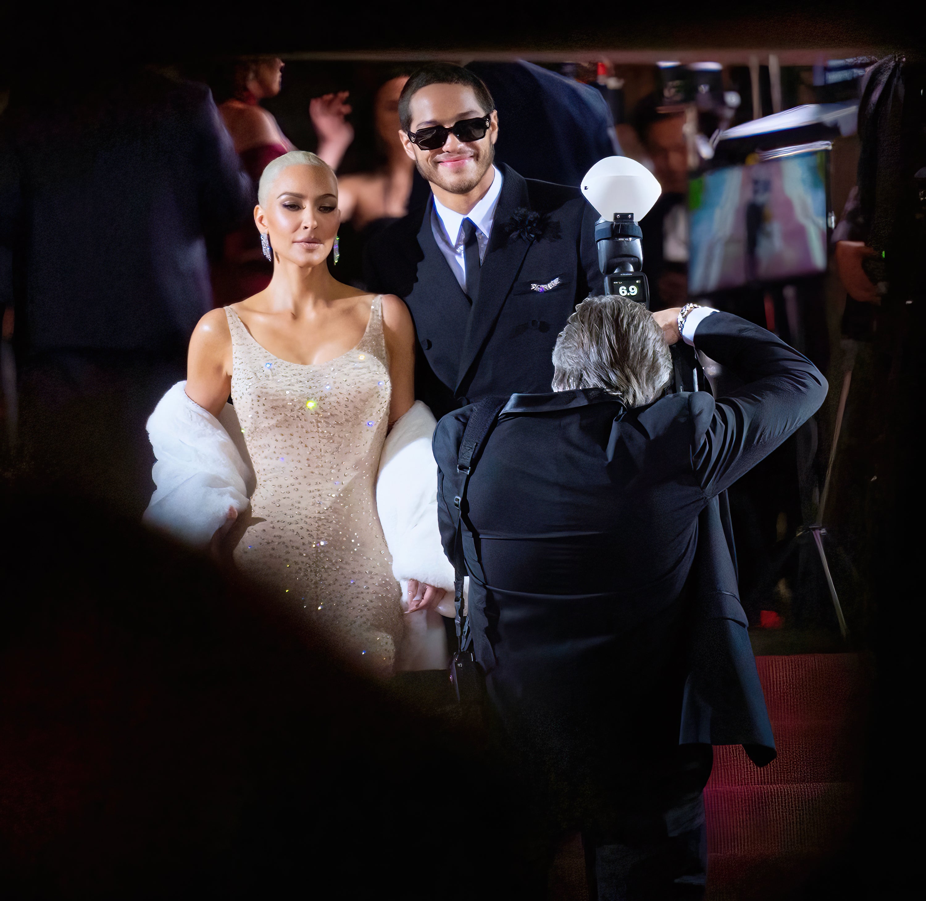 Two people pose on a red carpet; one in a sparkling gown with a stole, the other in a suit with sunglasses. Photographers capture the moment