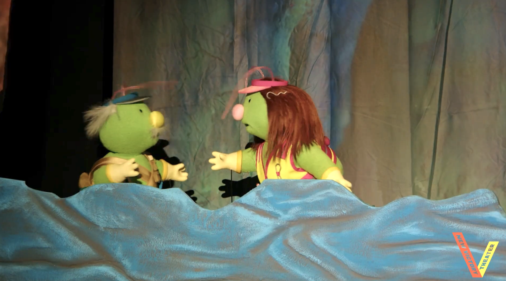 Two puppets with greenish faces and exaggerated features performing connected stage, interacting with each other