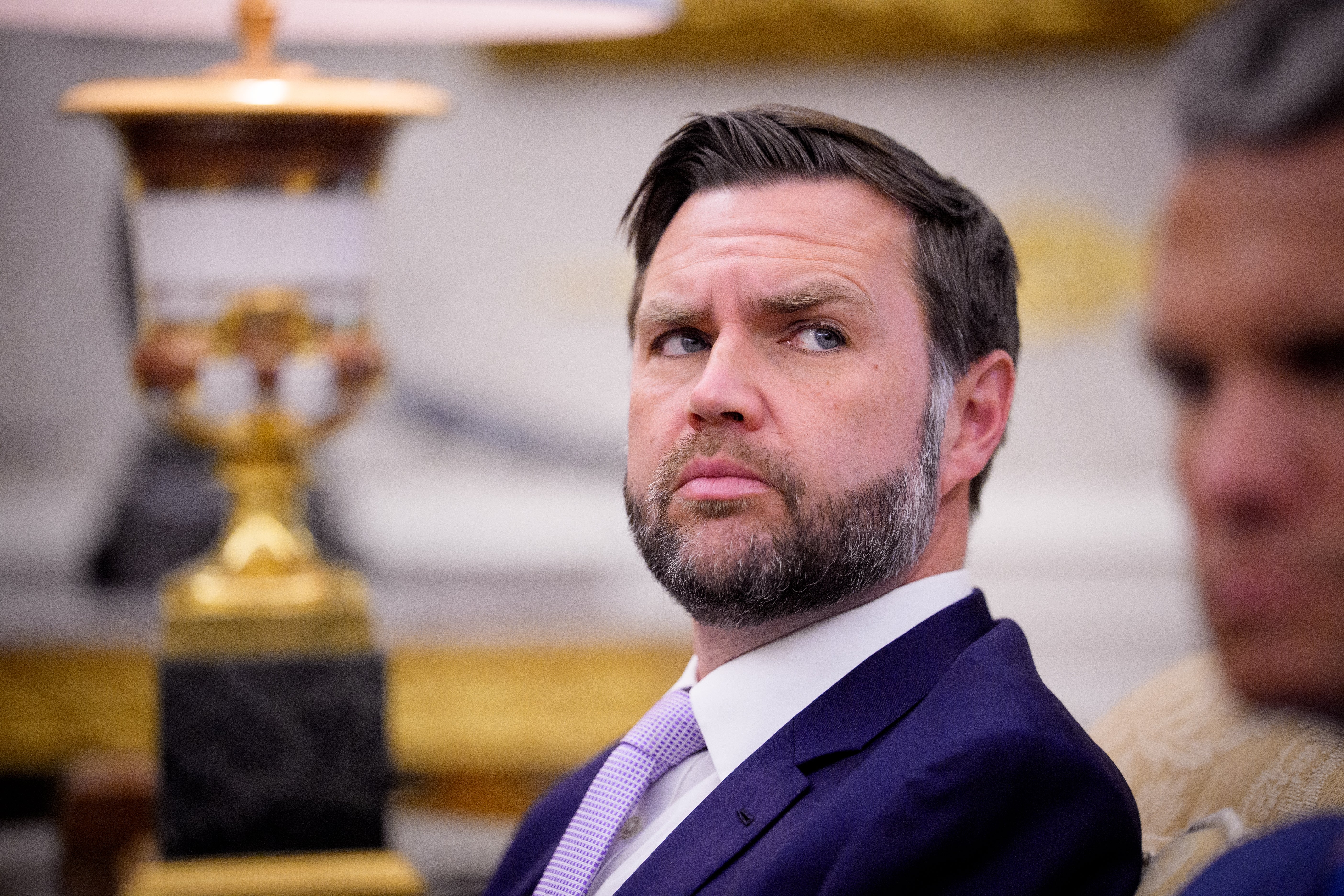 Person i formell antrekk med et gjennomtenkt uttrykk, sittende innendørs, med uskarpe figurer og en lampe i bakgrunnen