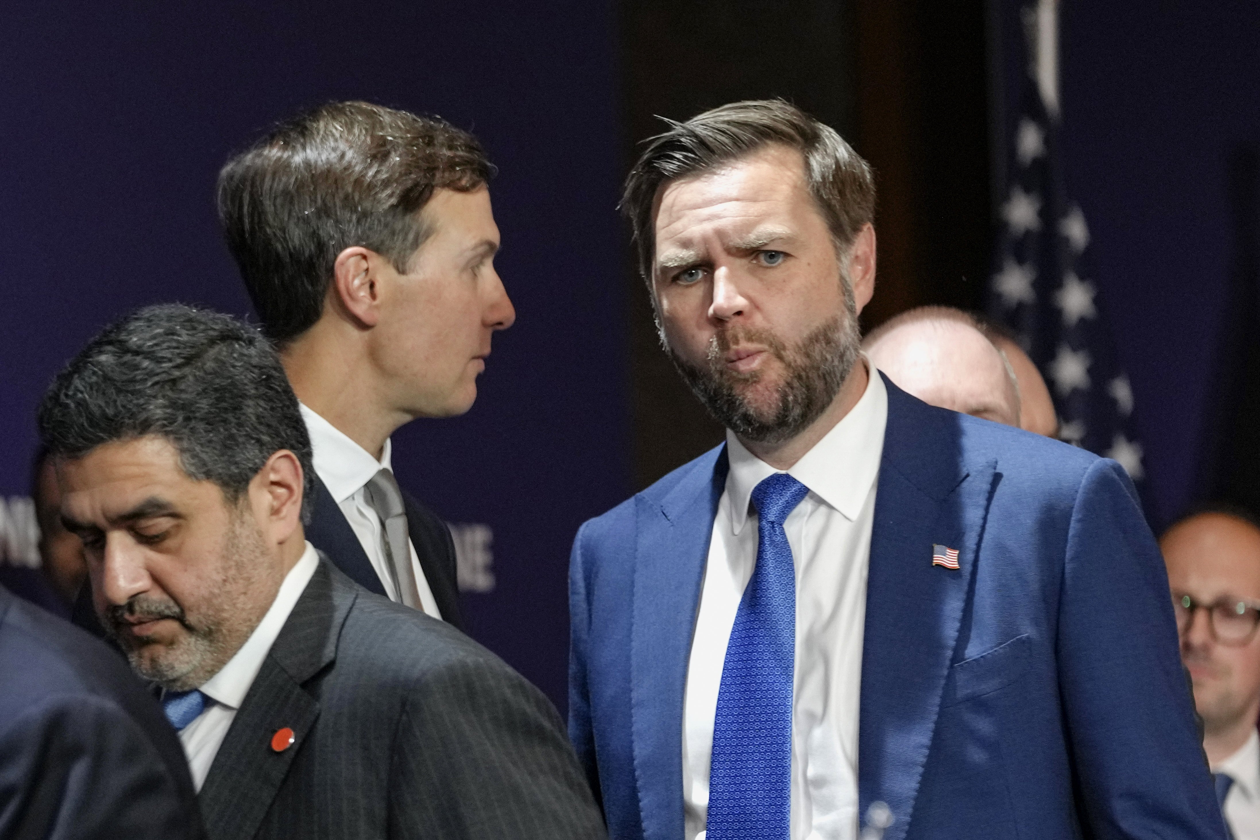 To menn i forretningsdress, engasjert i samtale ved en formell begivenhet. USAs flagg synlig i bakgrunnen