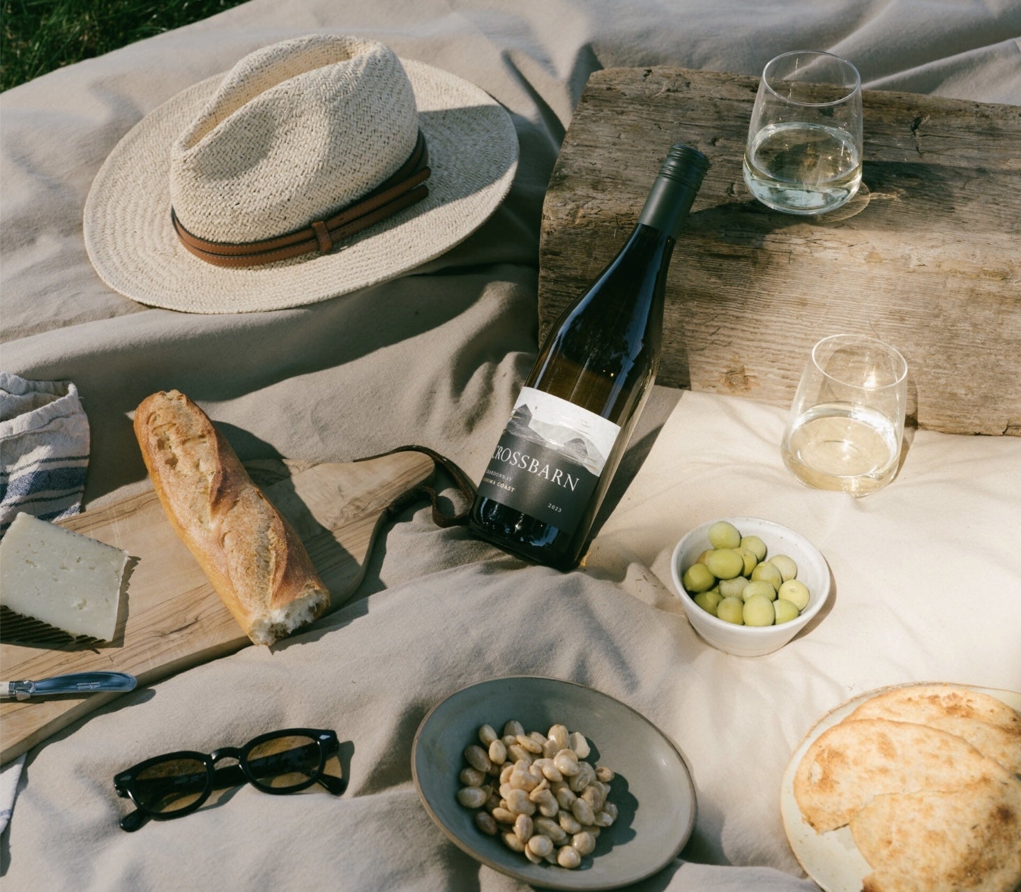 Picnic setup with wine, bread, cheese, greenish olives, nuts, sunglasses, and a straw chapeau connected a blanket