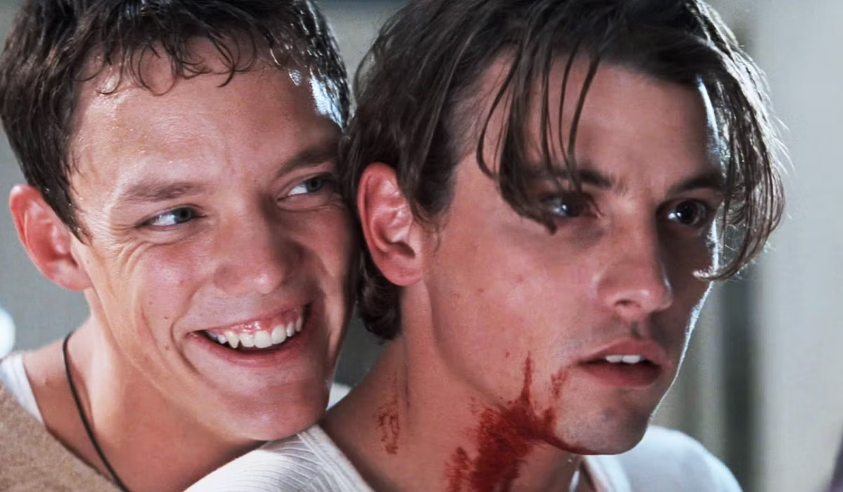 Two men appear in a tense movie scene; one looks distressed with fake blood, the other smiles behind him, hinting at a dramatic or horror film moment