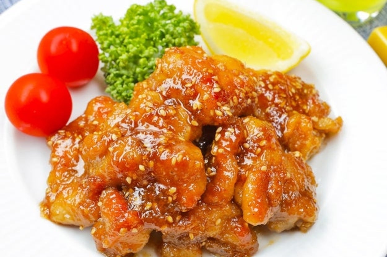 白い皿に盛り付けた鶏もも肉の甘辛ごま照り焼き。レモン、ブロッコリー、ミニトマトが添えられている。
