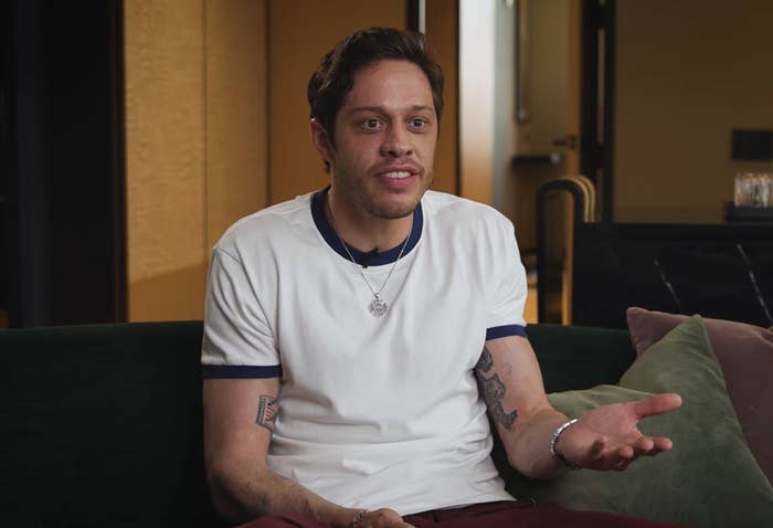 A idiosyncratic   with casually styled tattoos sits connected  a couch, wearing a achromatic  T-shirt and gesturing with 1  hand, appearing to beryllium  engaged successful  conversation