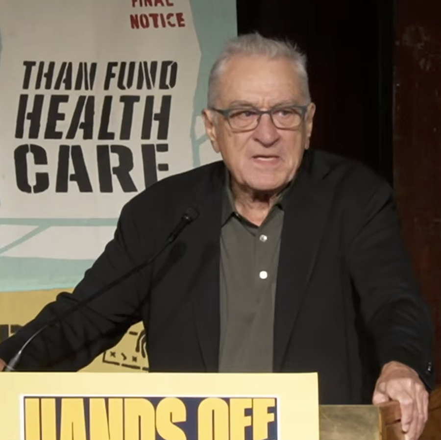 An older man speaks at a podium with a "Hands Off" sign, discussing healthcare advocacy