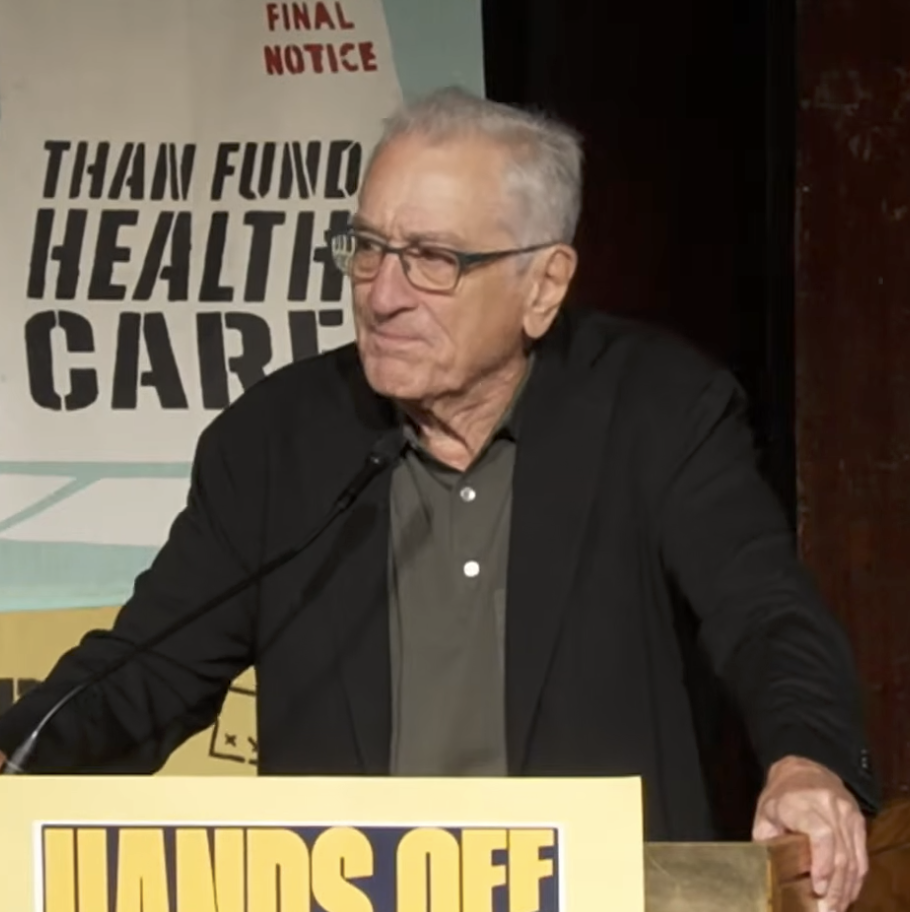 Robert De Niro speaks at a podium with a "Hands Off" sign, possibly discussing healthcare funding