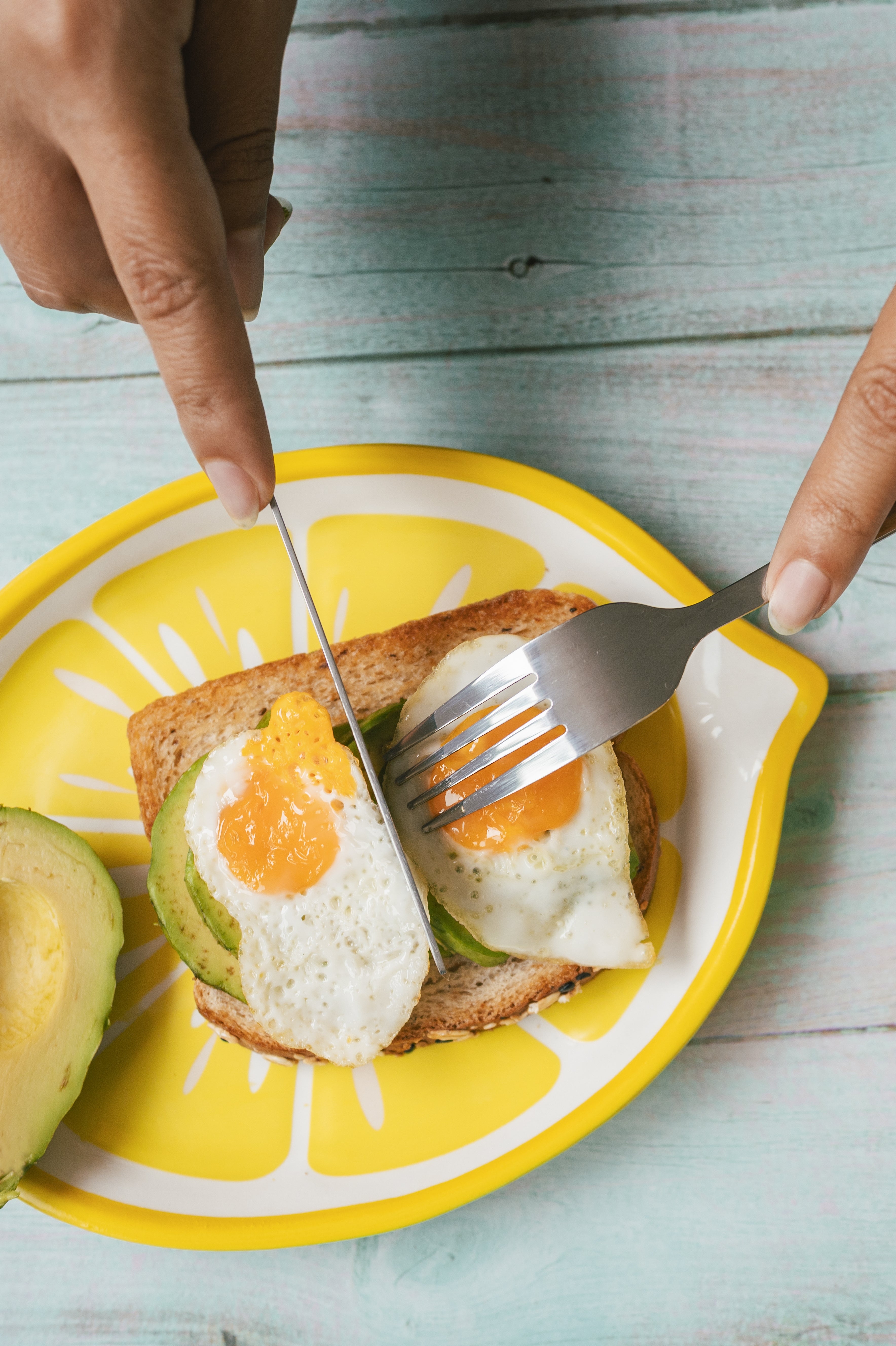 Person slicing avocado toast topped with a fried egg, using a fork and knife on a decorative plate