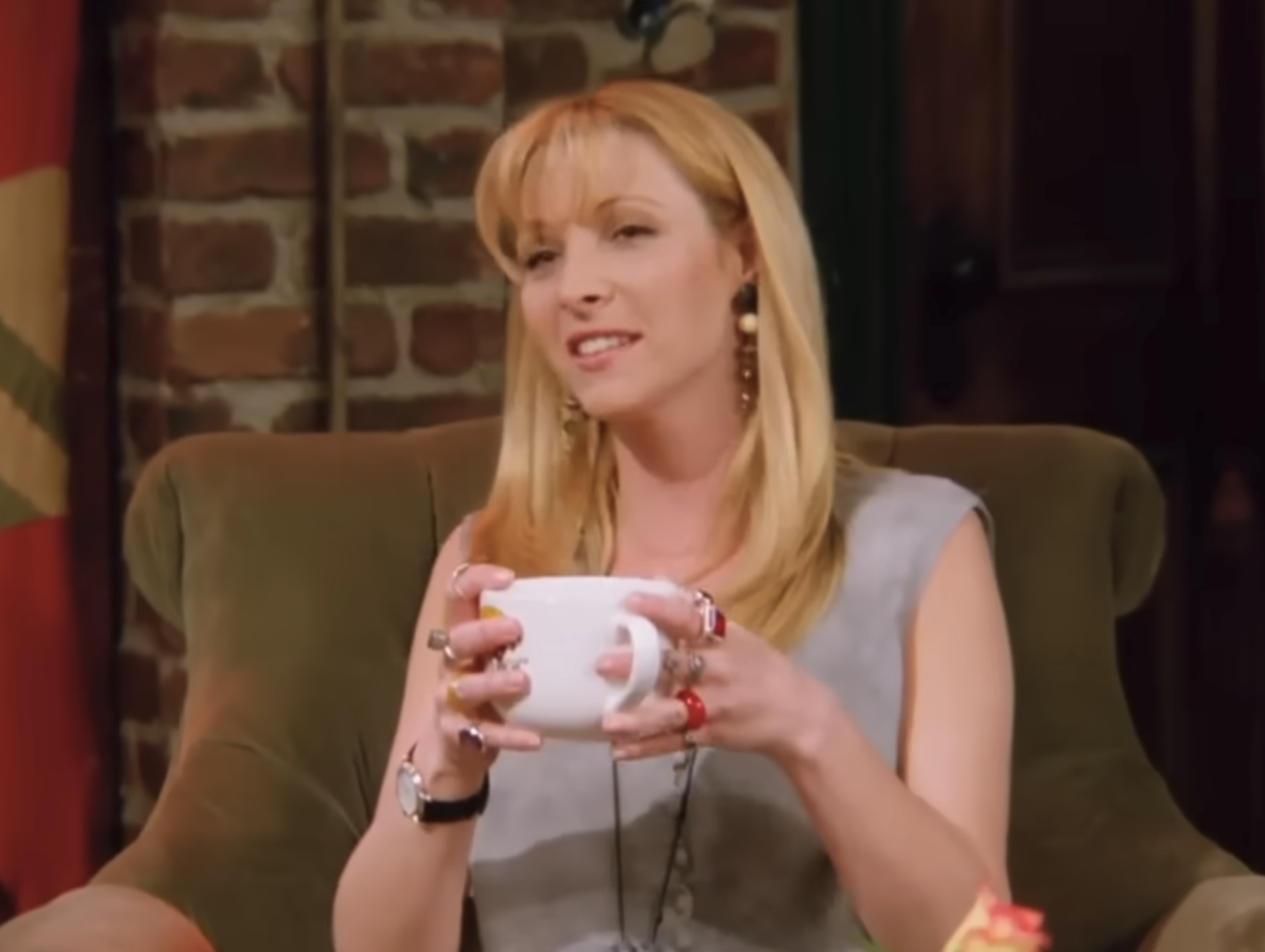 Person sitting on a sofa, holding a large mug, wearing casual attire, layered necklaces, and multiple rings, with a relaxed expression