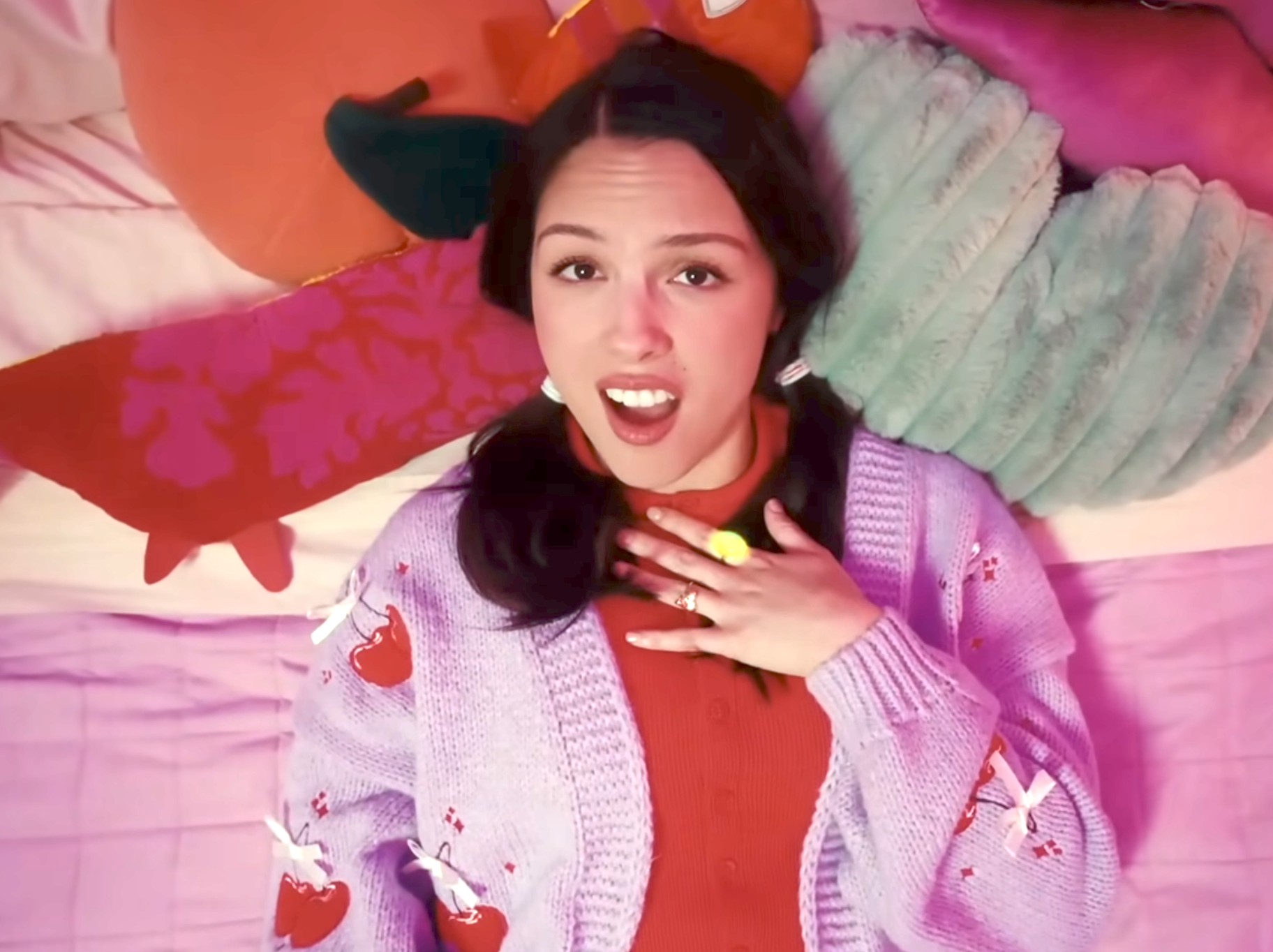 Person lying on a bed, surprised, wearing a patterned cardigan over a blouse, with playful pillows around