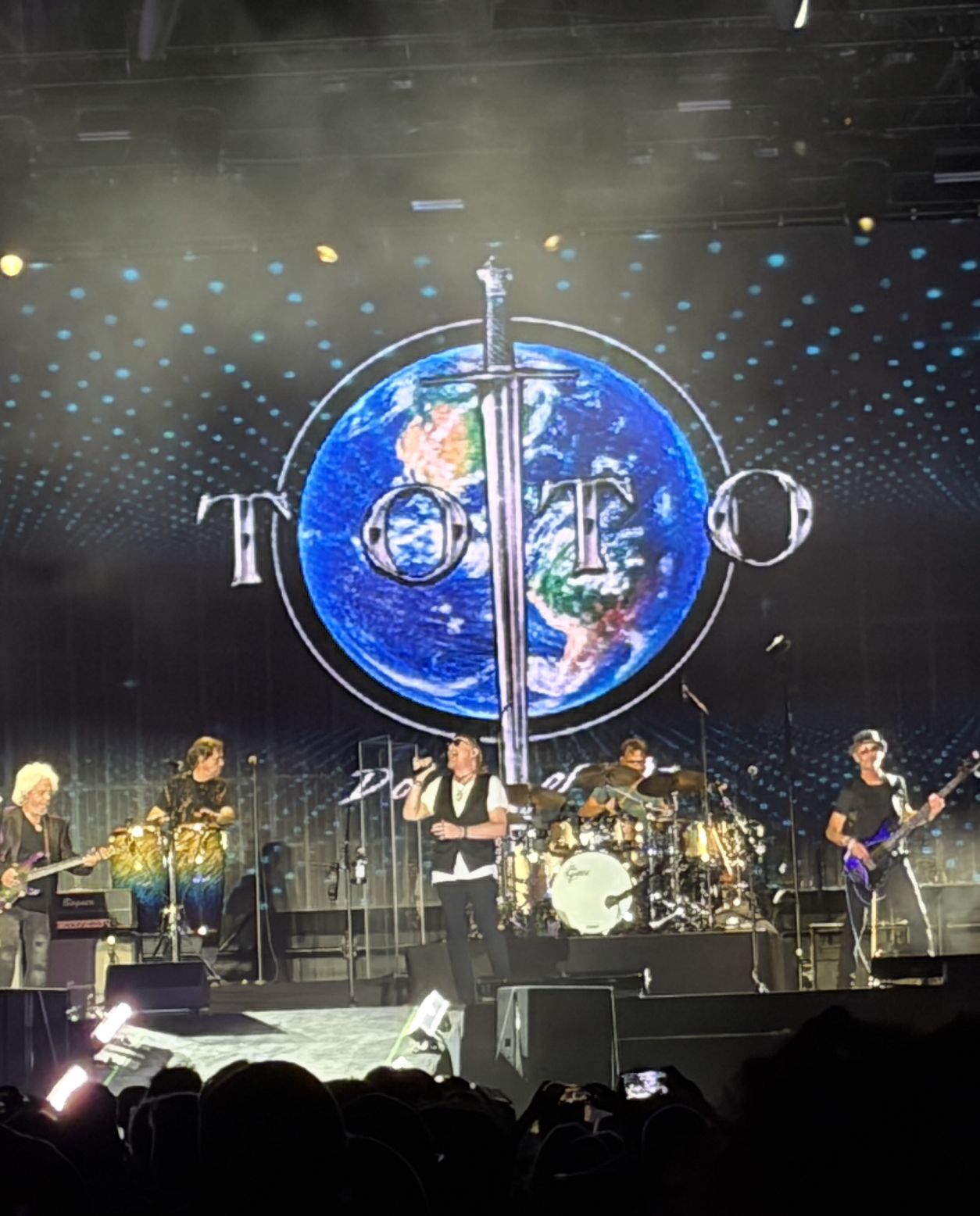 Band Toto performing connected signifier with Earth and sword logo successful the background. Members play instruments nether dynamic signifier lighting