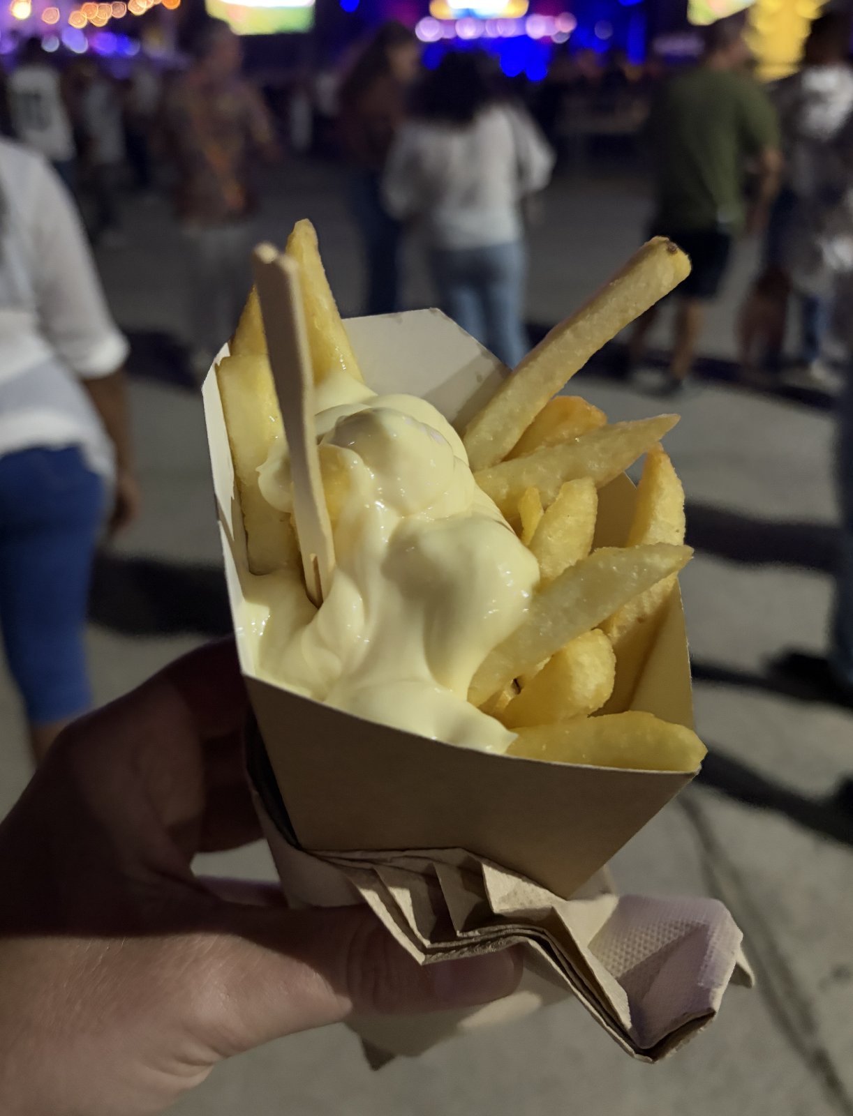 Hand holding a instrumentality of fries topped with a creamy condiment astatine a nighttime outdoor event. People and lights are blurred successful the background