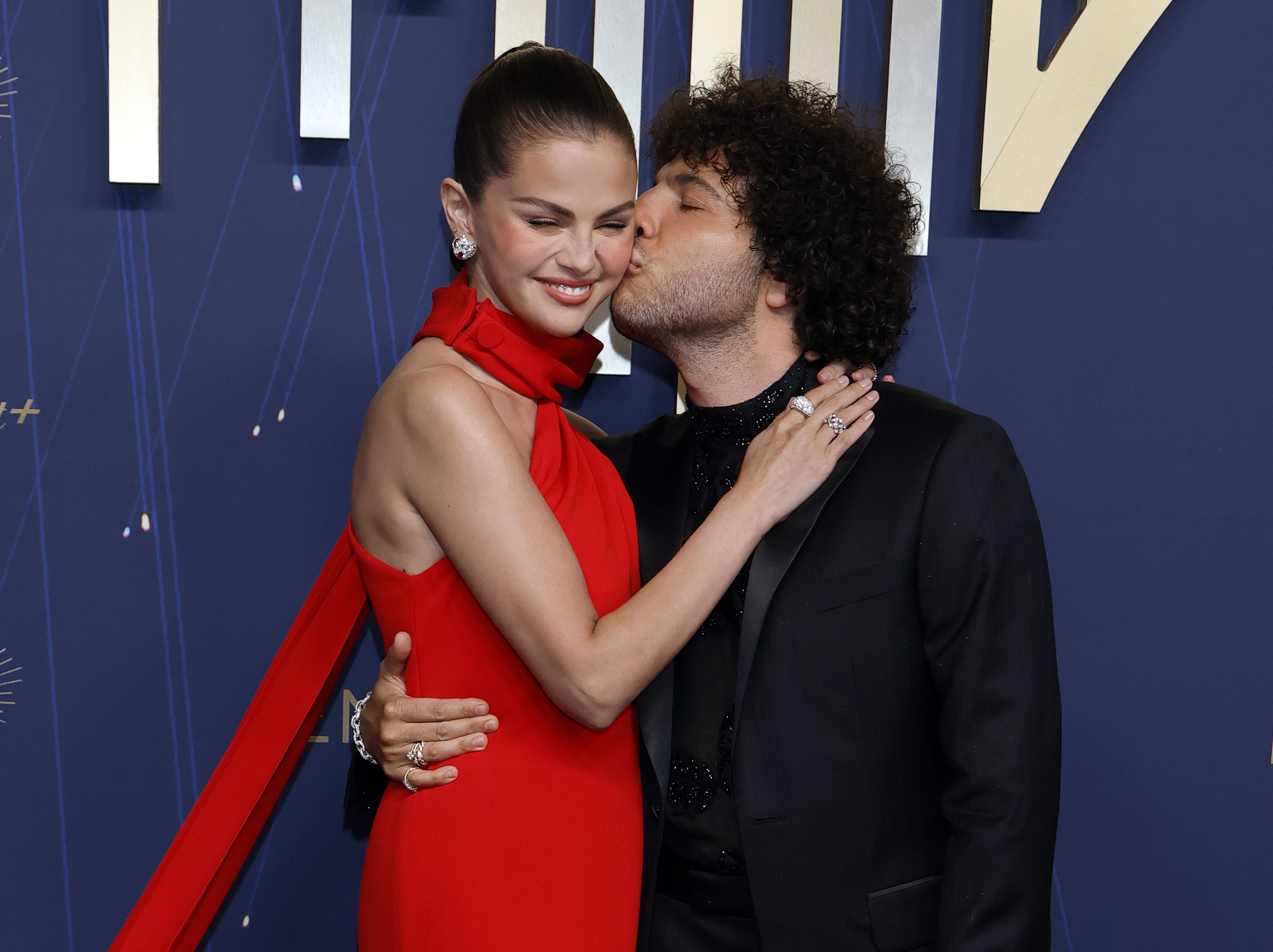 Selena Gomez in a sleek red dress being hugged and kissed on the cheek by Benny Blanco