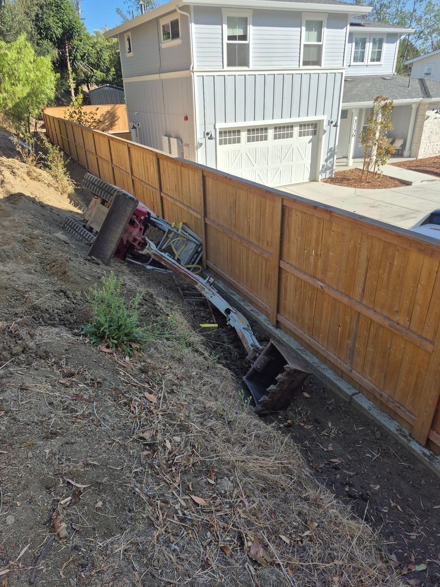 Bulldozer overturned on a residential slope near a wooden fence, showing a construction mishap in a suburban area