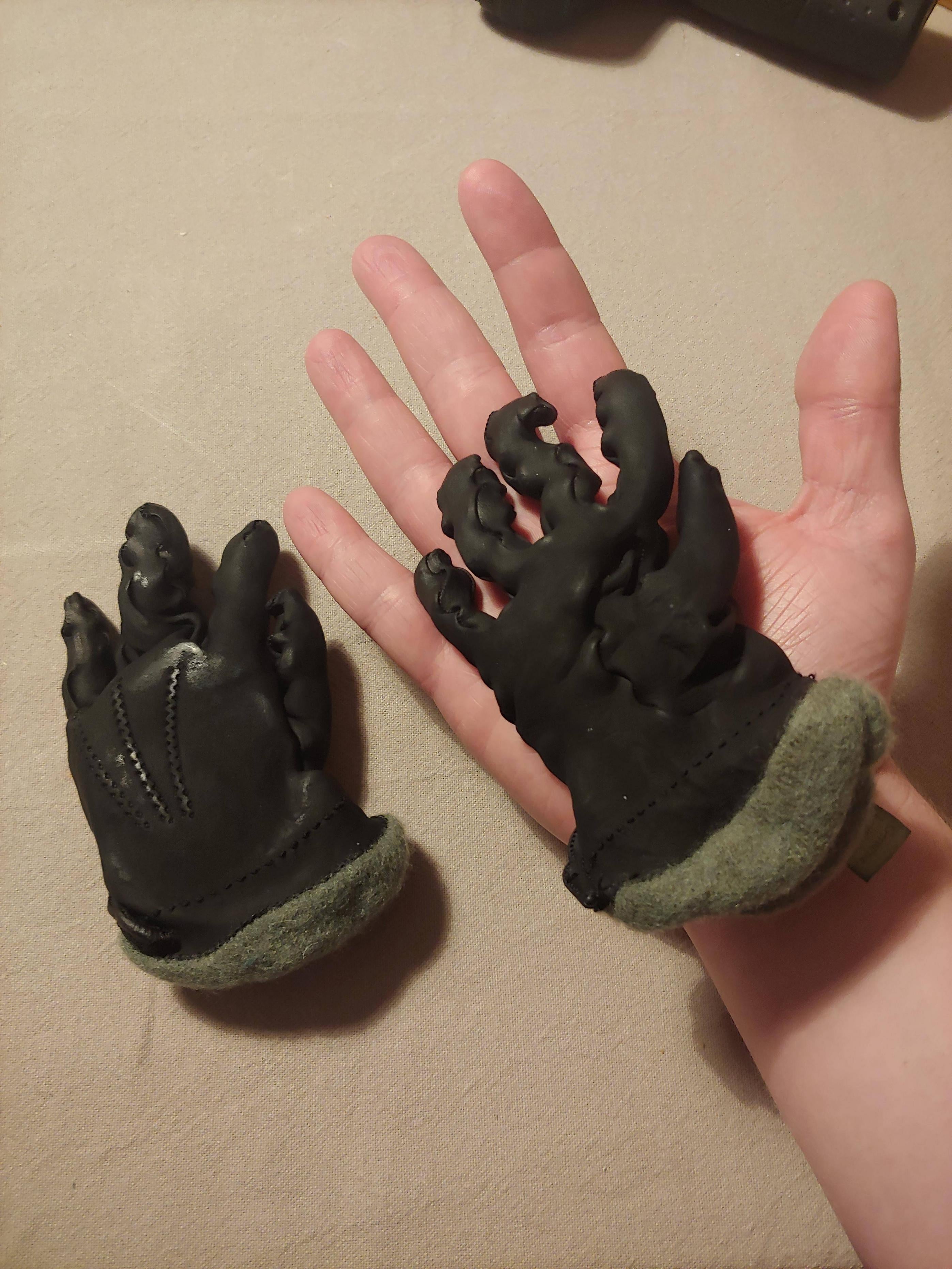 A hand holds a pair of detailed, small dragon claw gloves with fleece cuffs, showcasing the intricate design and texture