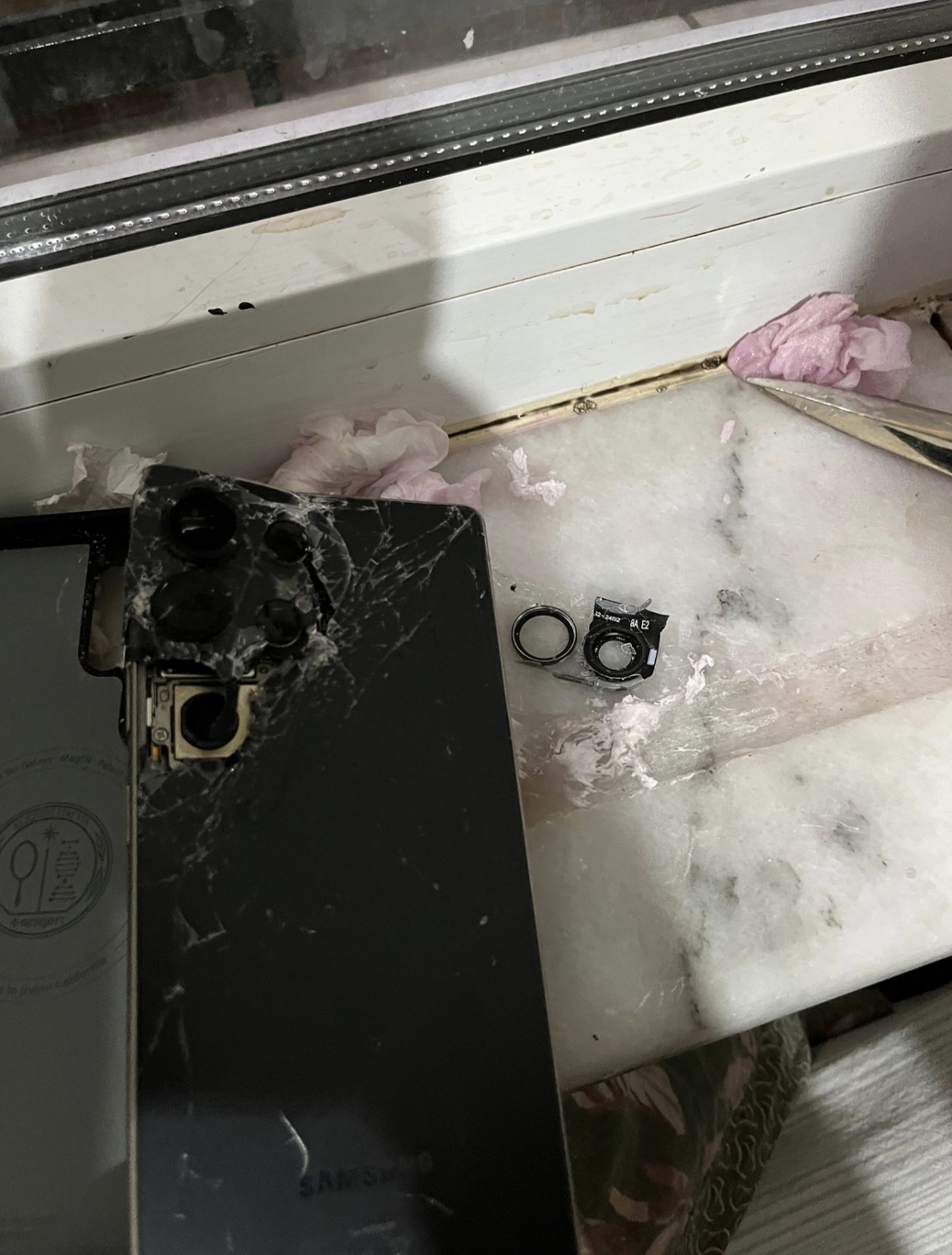 A broken phone with a cracked back and detached camera lenses on a windowsill