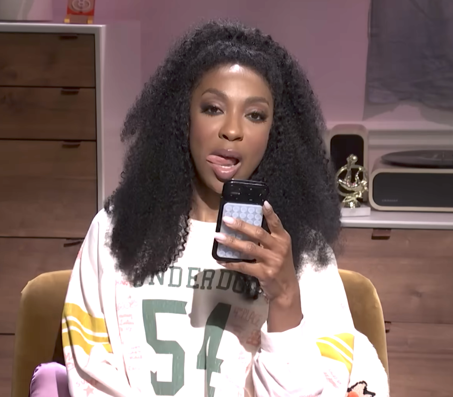 Person with curly hairsbreadth  sitting connected  a couch, playfully holding a telephone  to their mouth. They are wearing a casual printed sweatshirt