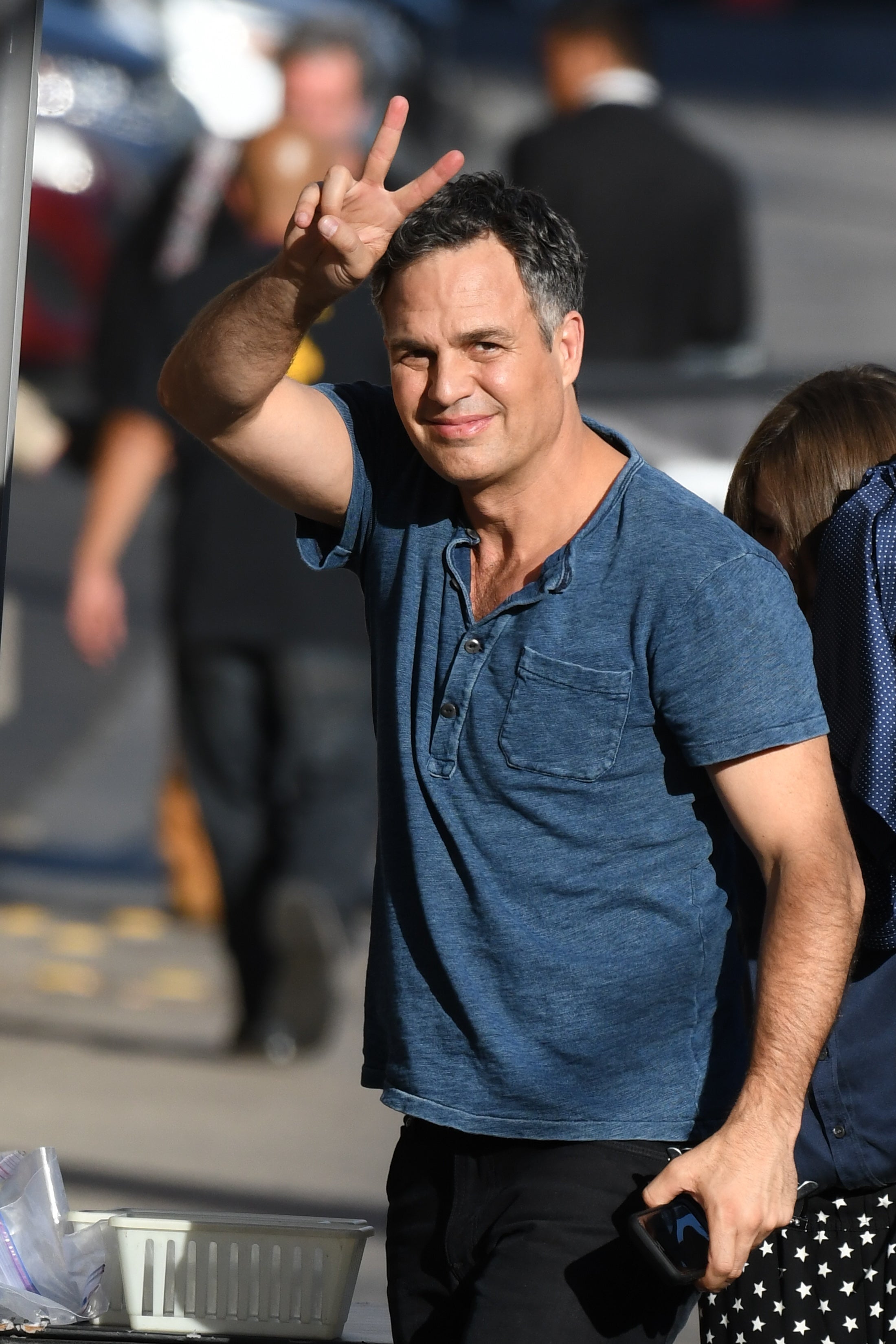 Mark Ruffalo in casual attire, making a peace sign with hand. They are smiling and walking