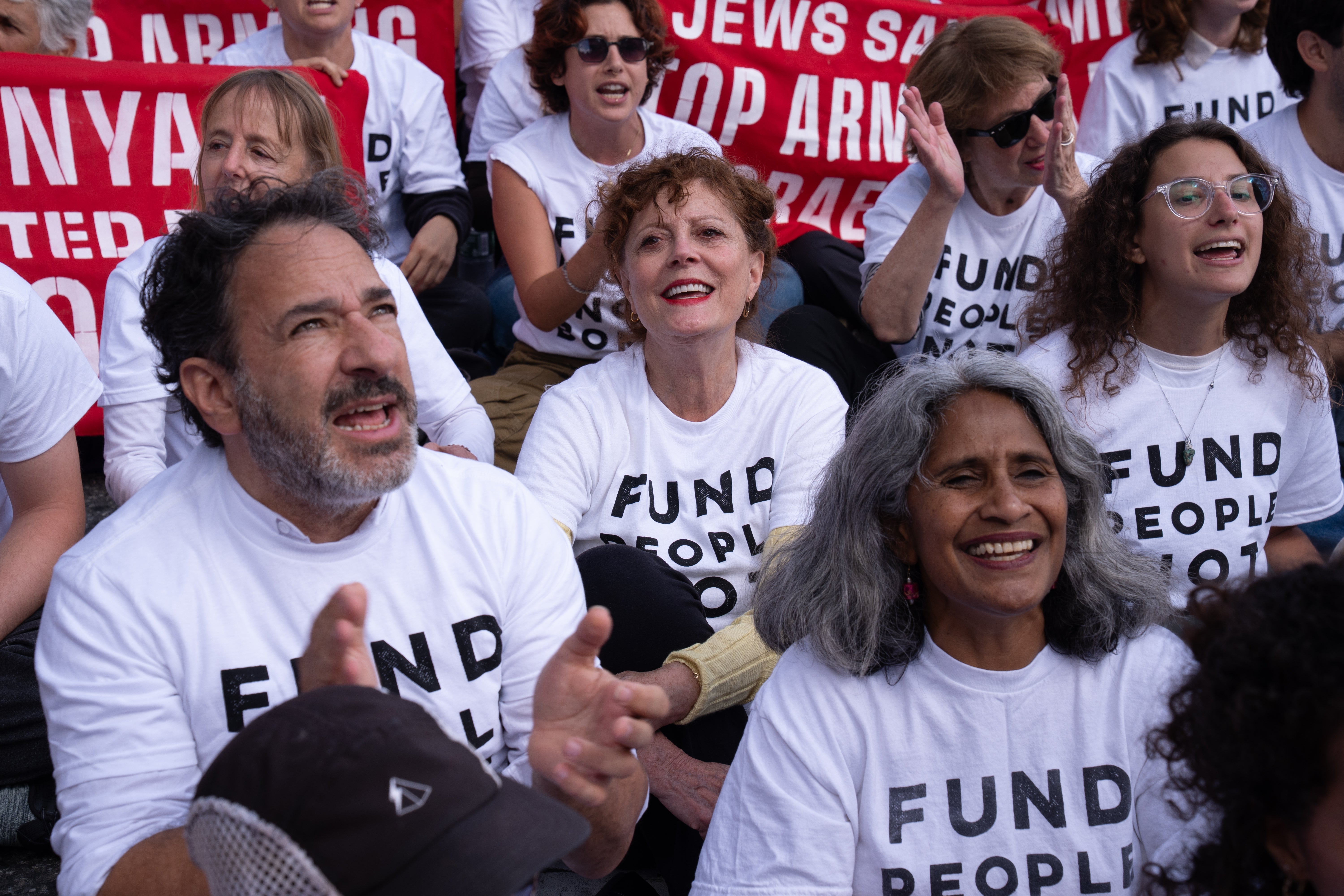 Susan Sarandon with a group of people wearing "Fund People Not Bombs" shirts sitting and clapping at a rally
