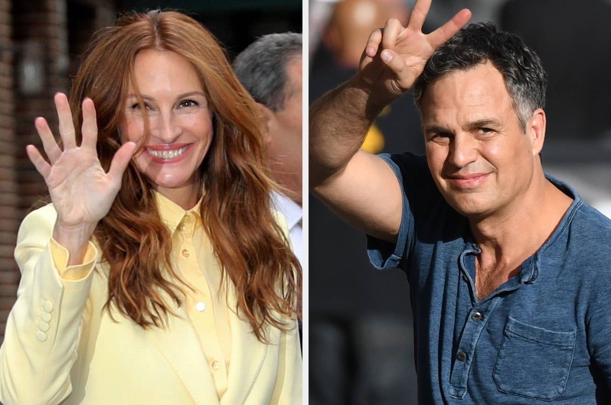 Two celebrities waving at an event; one is wearing a suit jacket over a buttoned shirt, and the other in a casual shirt with a subtle neckline