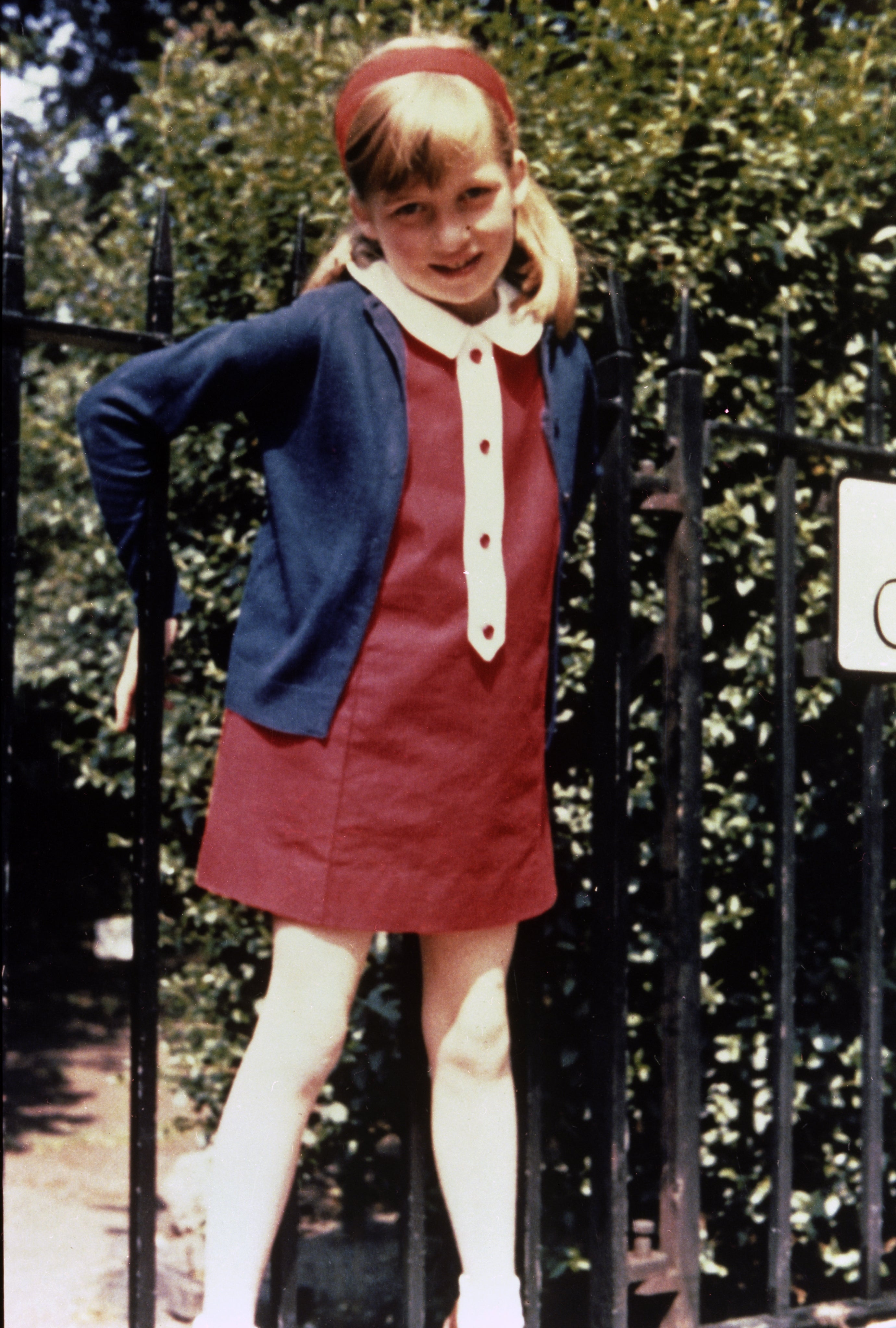 Lady Diana Spencer wearing a red dress, blue sweater while she stands by a fence