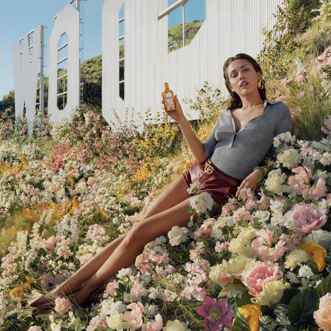 Person in stylish casual outfit reclines in a flower-filled landscape, holding a skincare product with a large sign in the background
