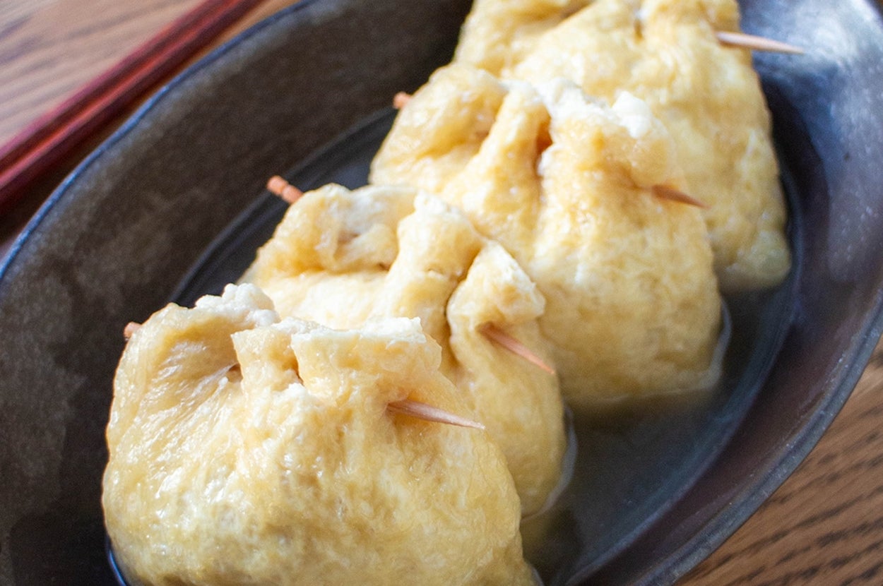 お皿に盛られた三つの巾着、油揚げで包まれた料理。箸が横に置かれている。