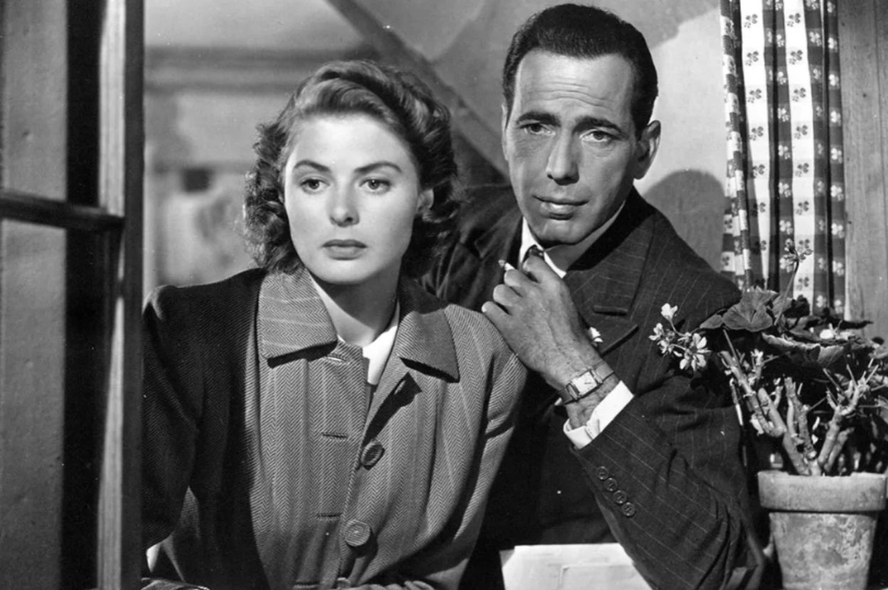 Two actors from a classic film scene, one in a pinstripe suit, the other in a collared coat, sit closely by a window with thoughtful expressions