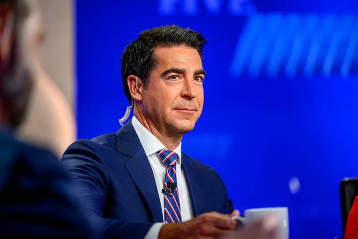 Man in suit with tie, wearing an earpiece, sits at a table on a TV set, suggesting participation in a broadcast or interview setting