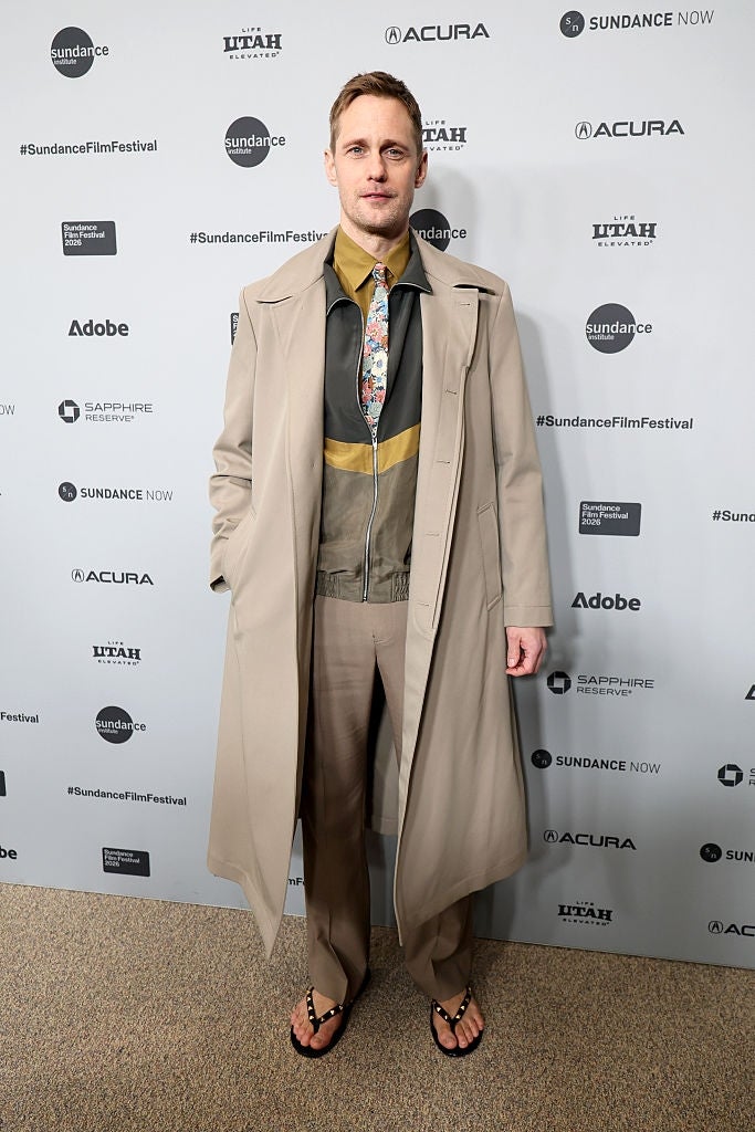 Person on red carpet wearing a long coat, patterned shirt, tie, and sandals at Sundance Film Festival