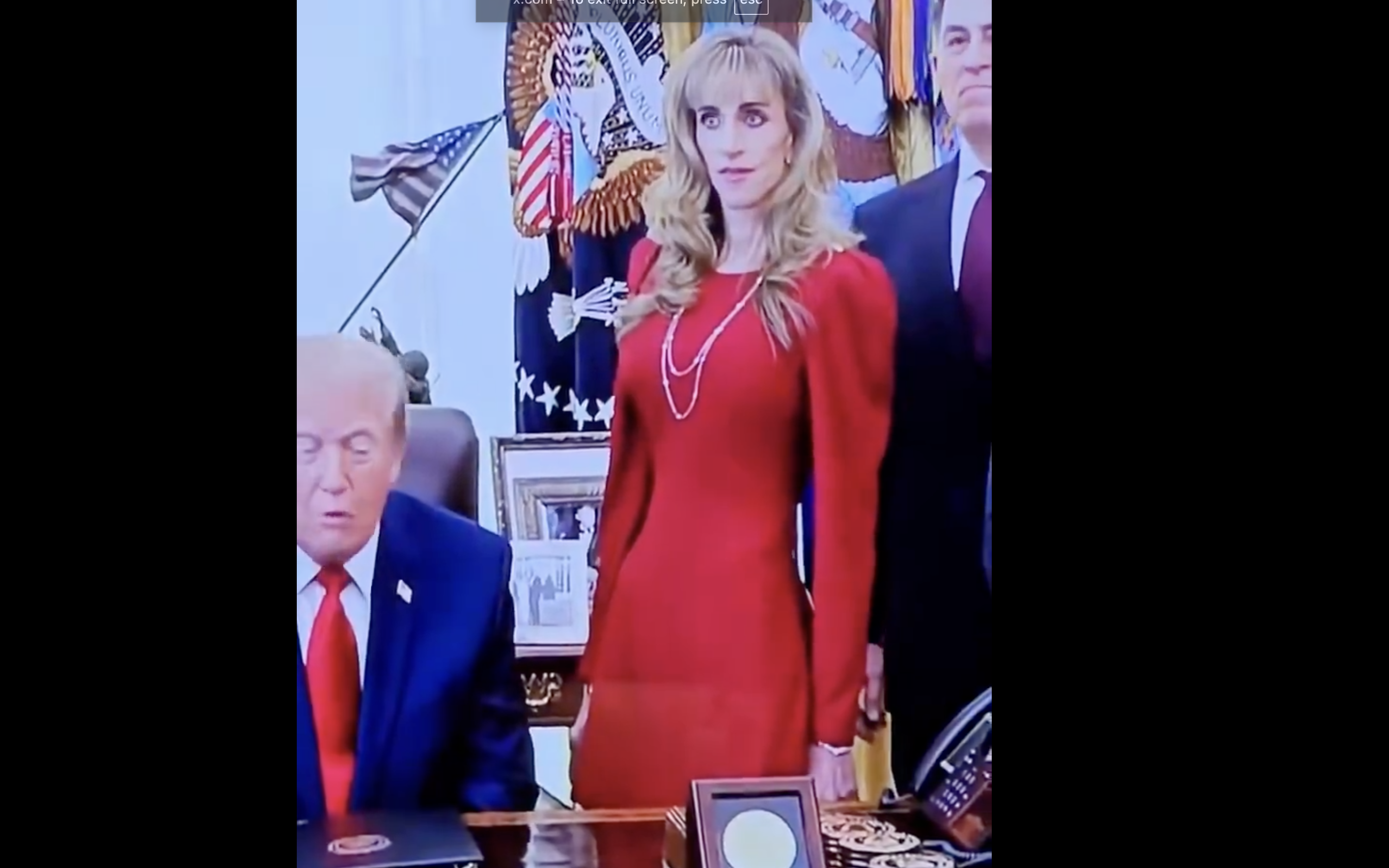 A person in a red dress stands beside a seated individual in a suit and red tie, in an official setting with flags and framed items