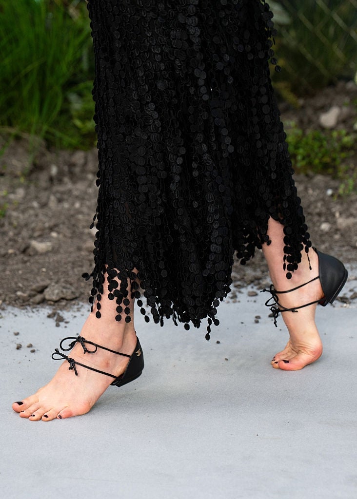 Barefoot individual walking on a gray surface, wearing a dress with a textured, dangling hem and lace-up flats