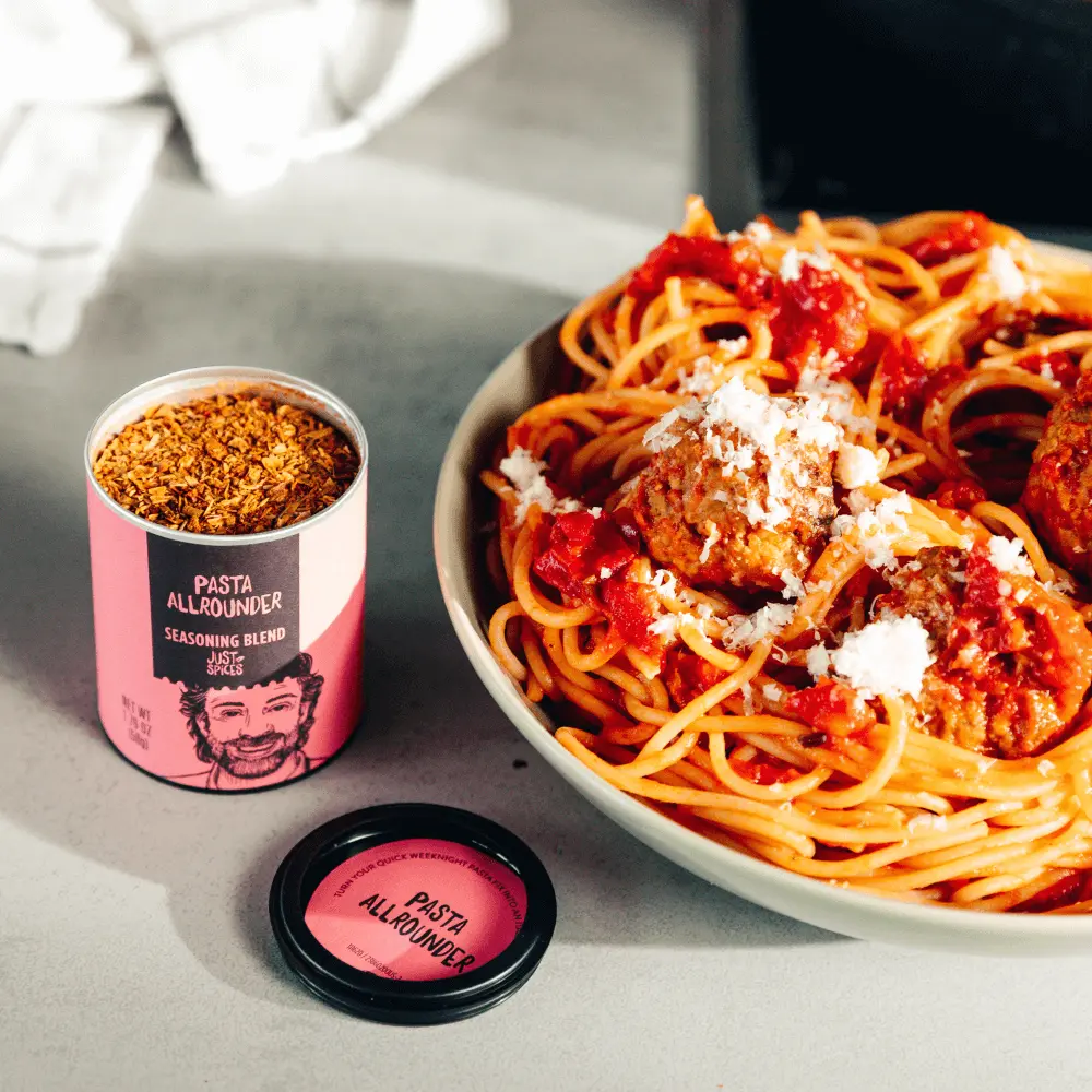 A plate of spaghetti with meatballs is next to an open spice container labeled "Pasta Allrounder Seasoning Blend."