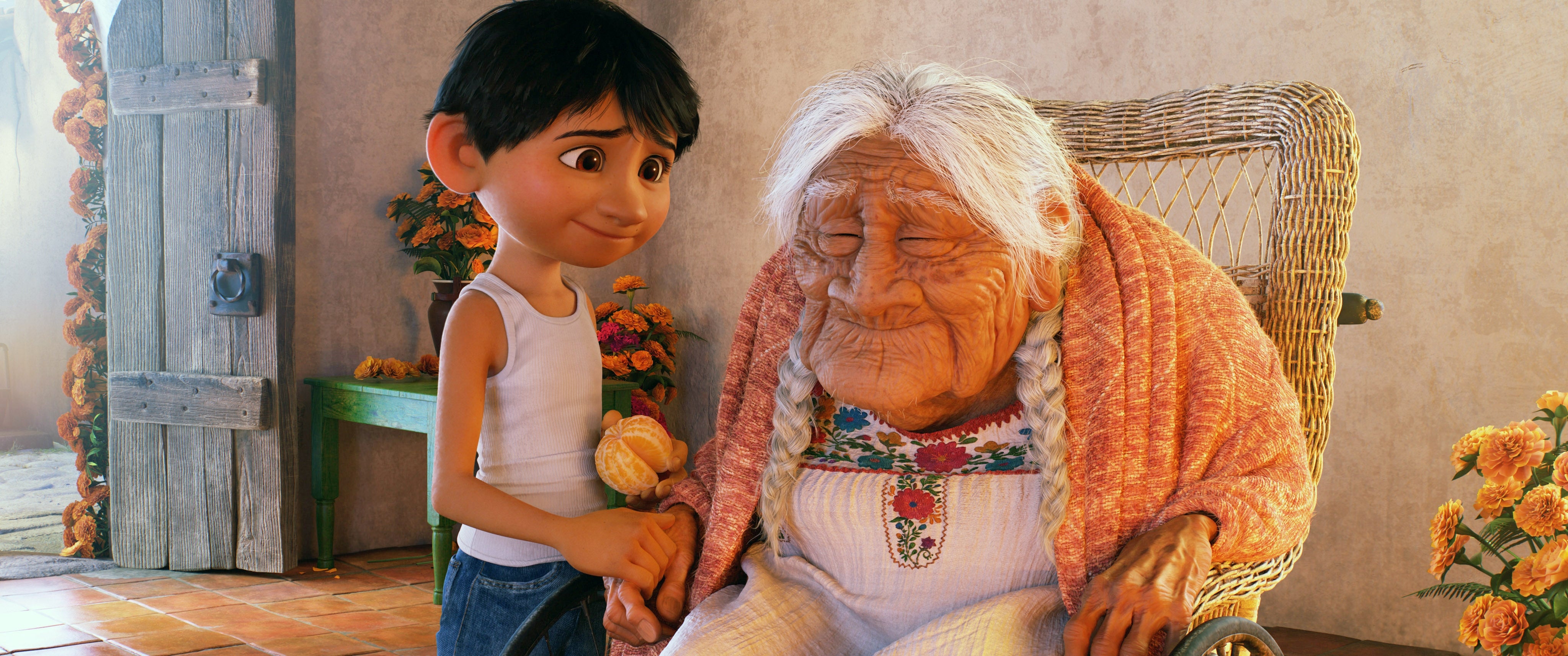 Miguel and mama coco in a cozy setting. Boy, in a tank top, holds her hand affectionately. Woman sits in a wicker chair, wearing a shawl