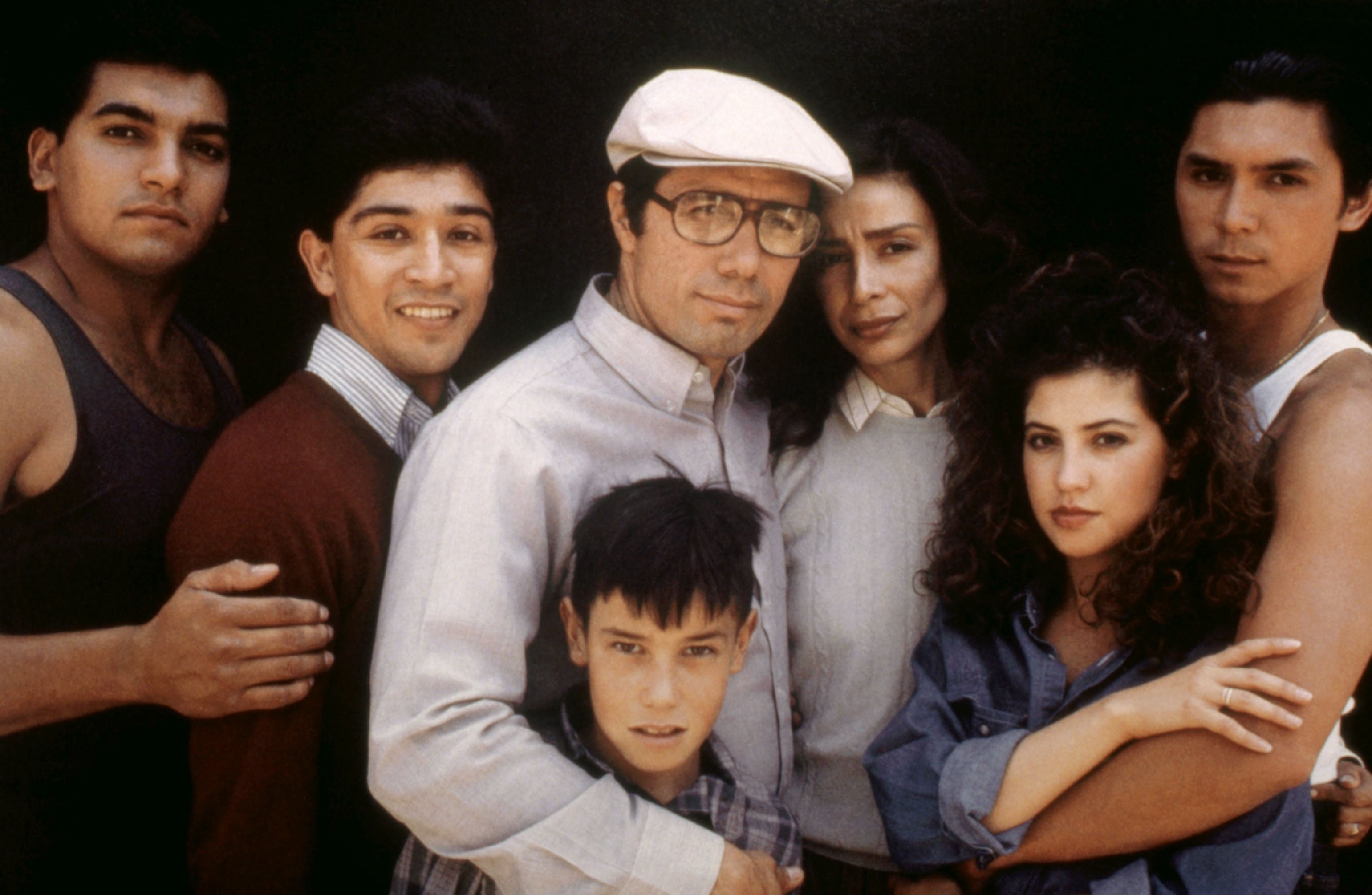 Group of seven people posing closely together with serious and thoughtful expressions. One person wears a cap and glasses. Casual attire