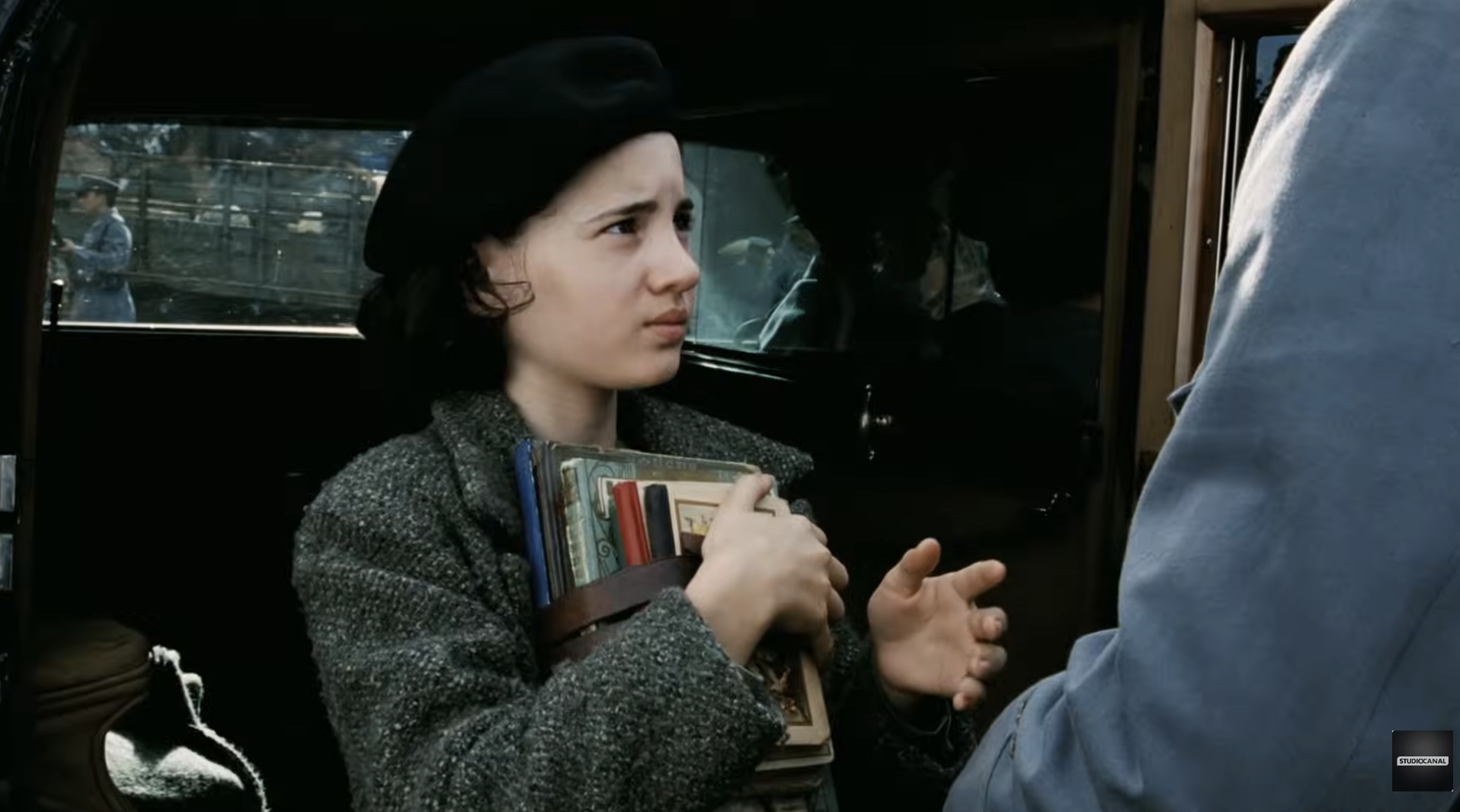 Young person in a beret and jacket, holding books, stands near a car, looking towards a figure in front of them