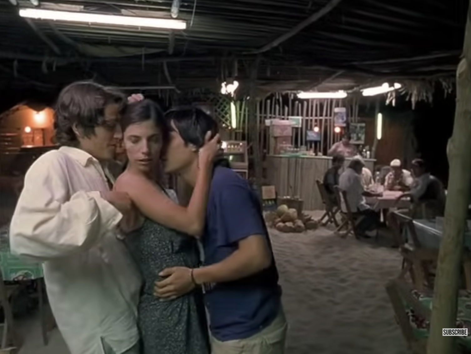 Three people embrace closely at an open-air cafe with sandy floors, while others dine in the background