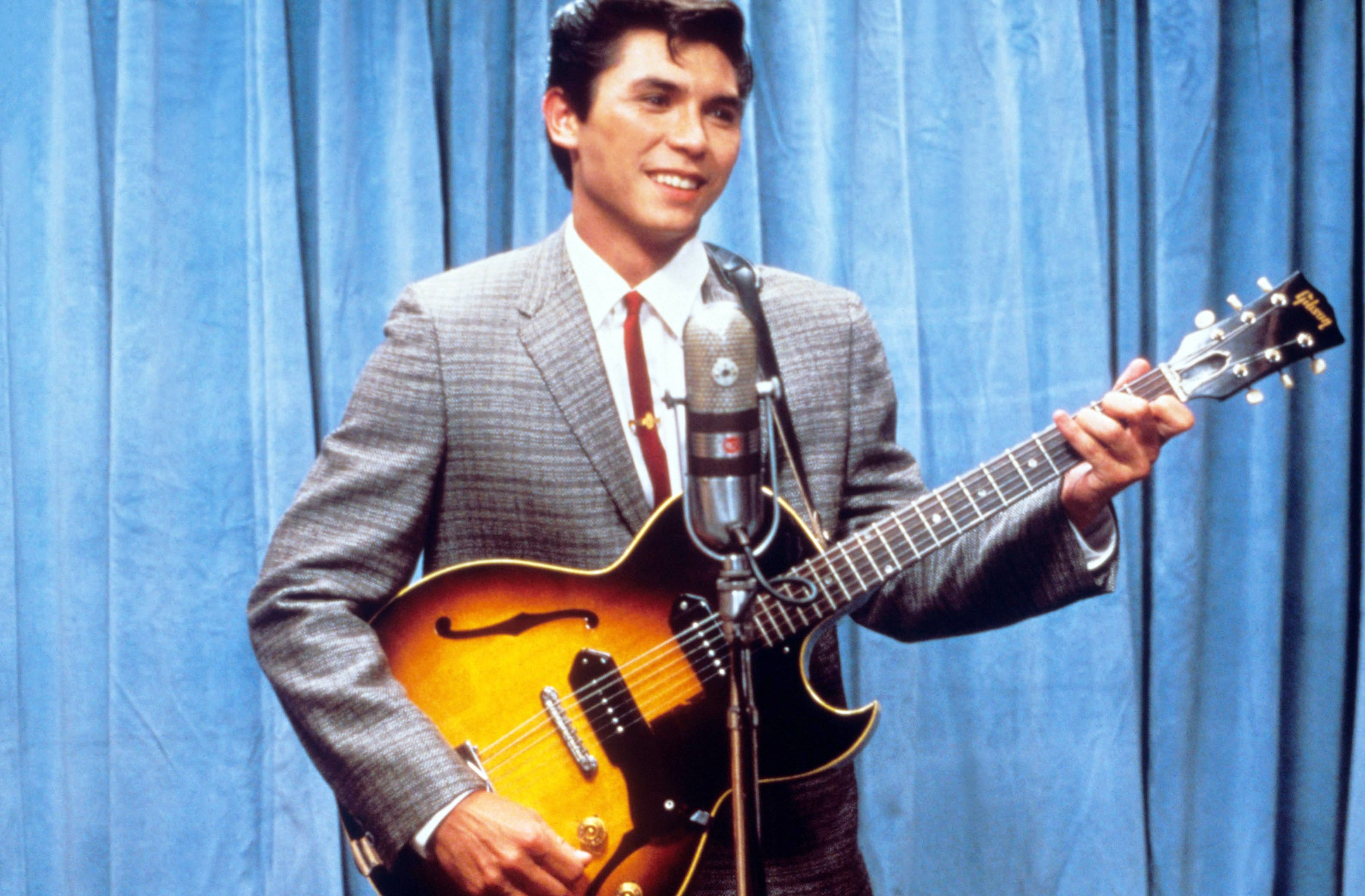 Person in a suit playing an electric guitar in front of a microphone, standing against a curtain backdrop